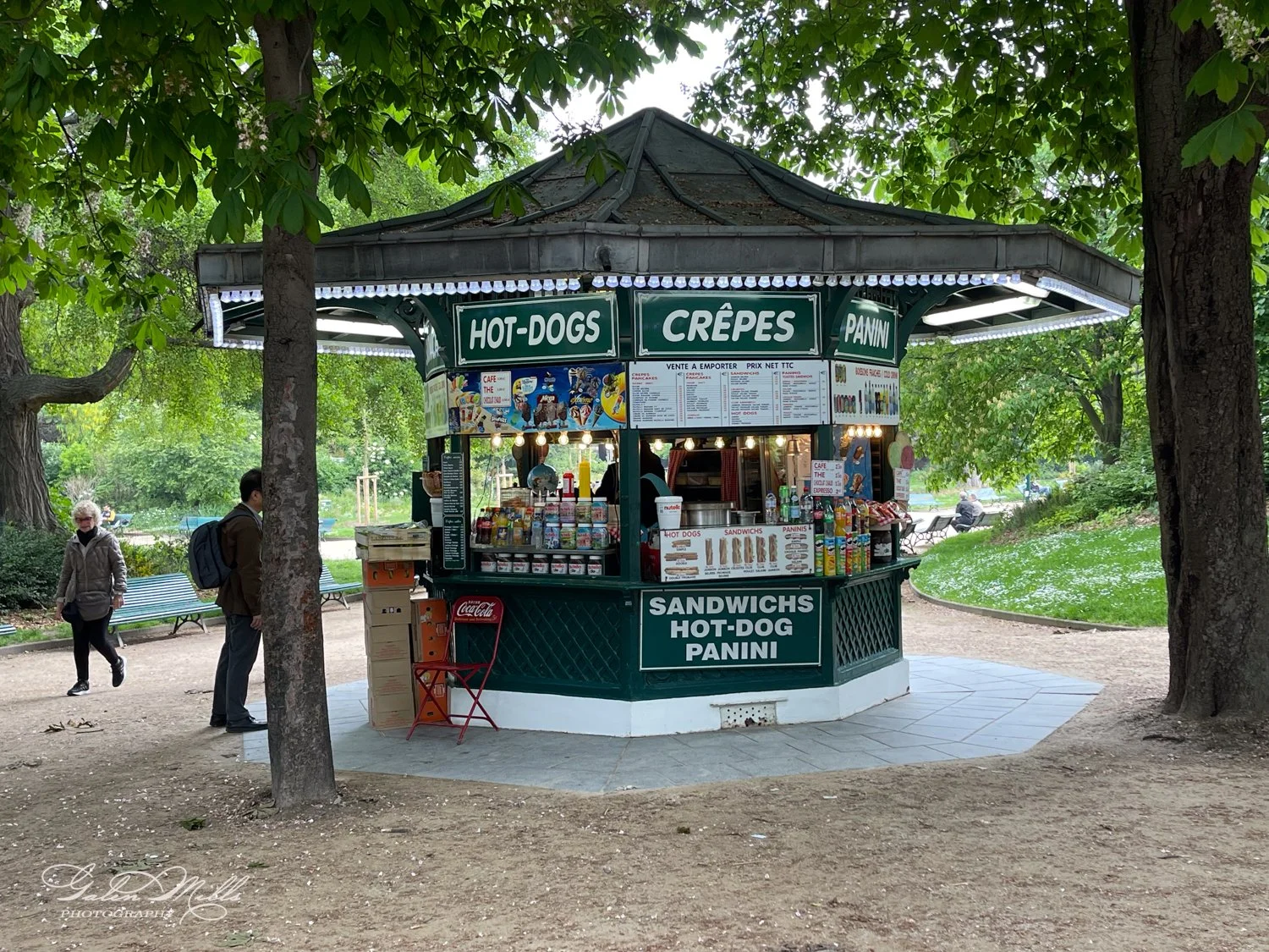 Park kiosk selling hot dogs, crepes, and panini, surrounded by greenery, with people walking by and park benches nearby.