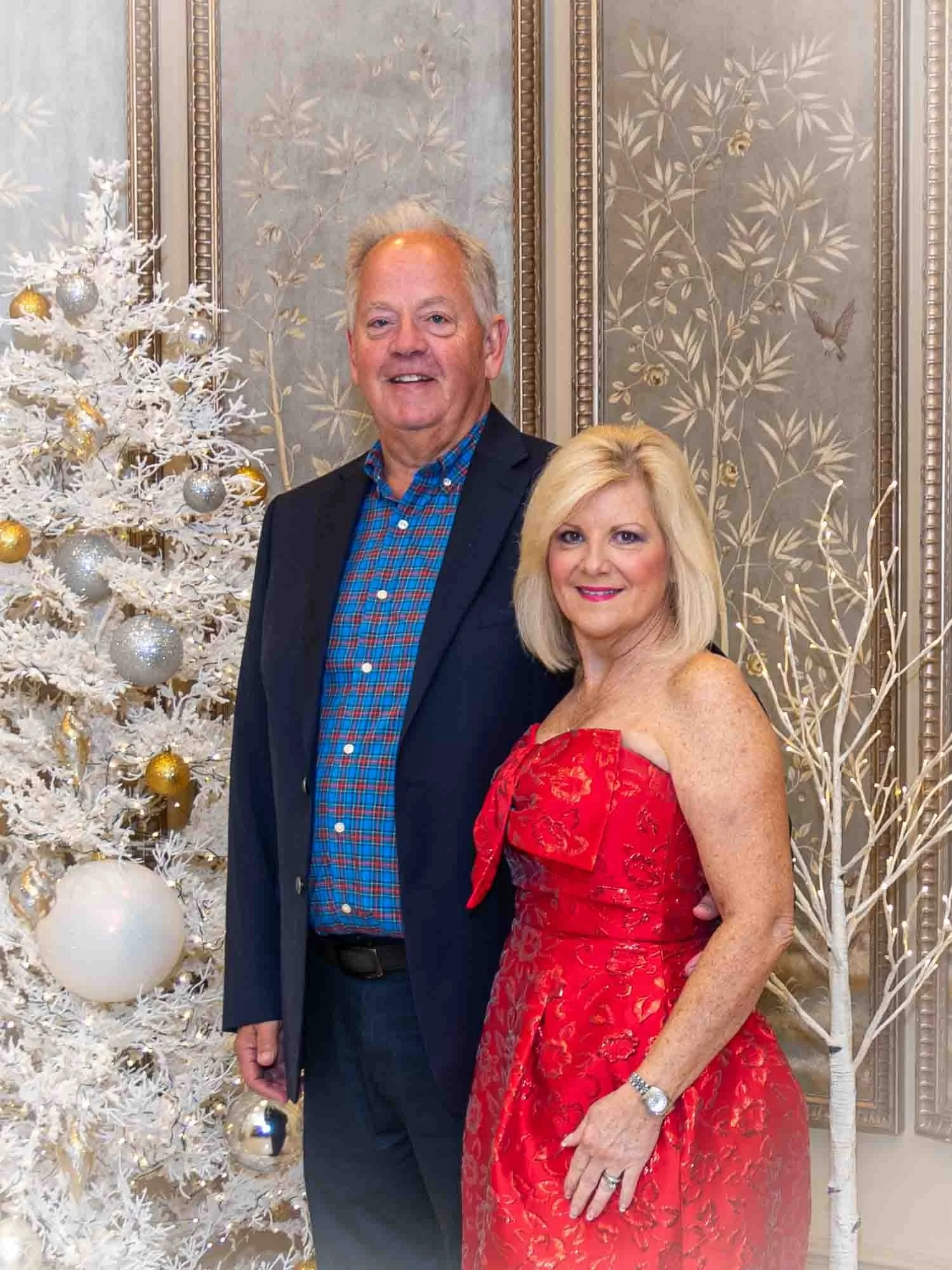 A man in a suit and a woman in a red dress posing in front of a white Christmas tree decorated with silver and gold ornaments. The background features ornate wallpaper with floral patterns.