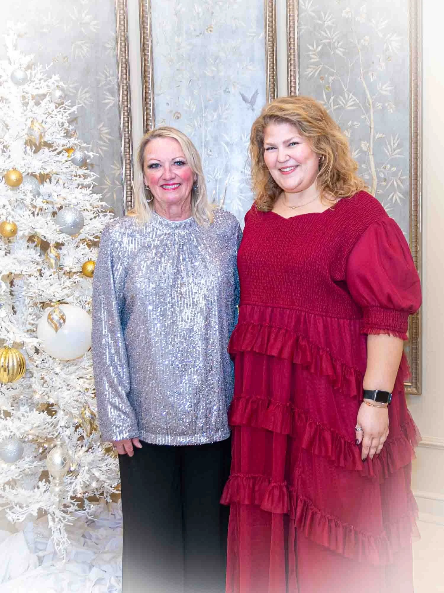 Two women pose in front of a white decorated Christmas tree with gold and silver ornaments. One wears a shimmering silver top and black pants, and the other wears a red ruffled dress.