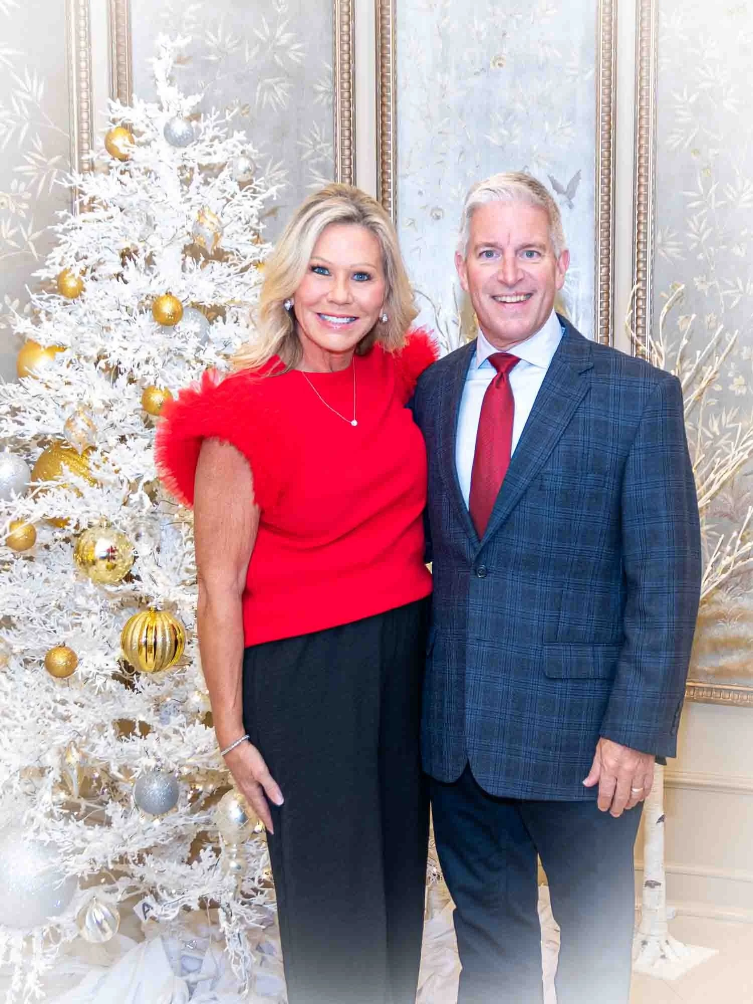 A man and woman posing in front of a white and gold decorated Christmas tree. The woman is wearing a red top and black pants, and the man is wearing a blue plaid suit with a red tie. The background includes ornate wallpaper and holiday decorations.