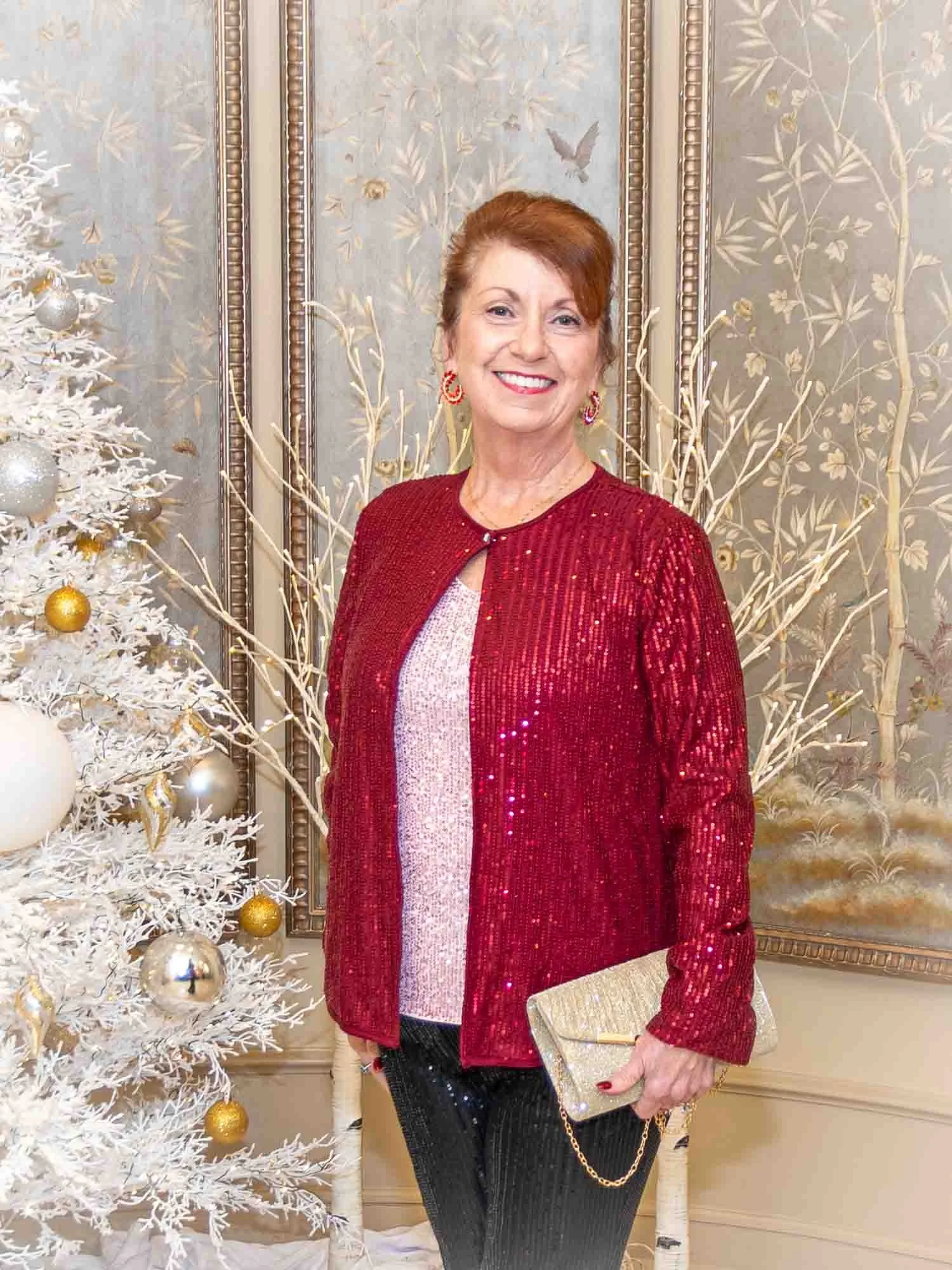 Woman in red sequin jacket holding a gold clutch, standing by a white decorated Christmas tree with gold and silver ornaments, against a floral patterned wall.