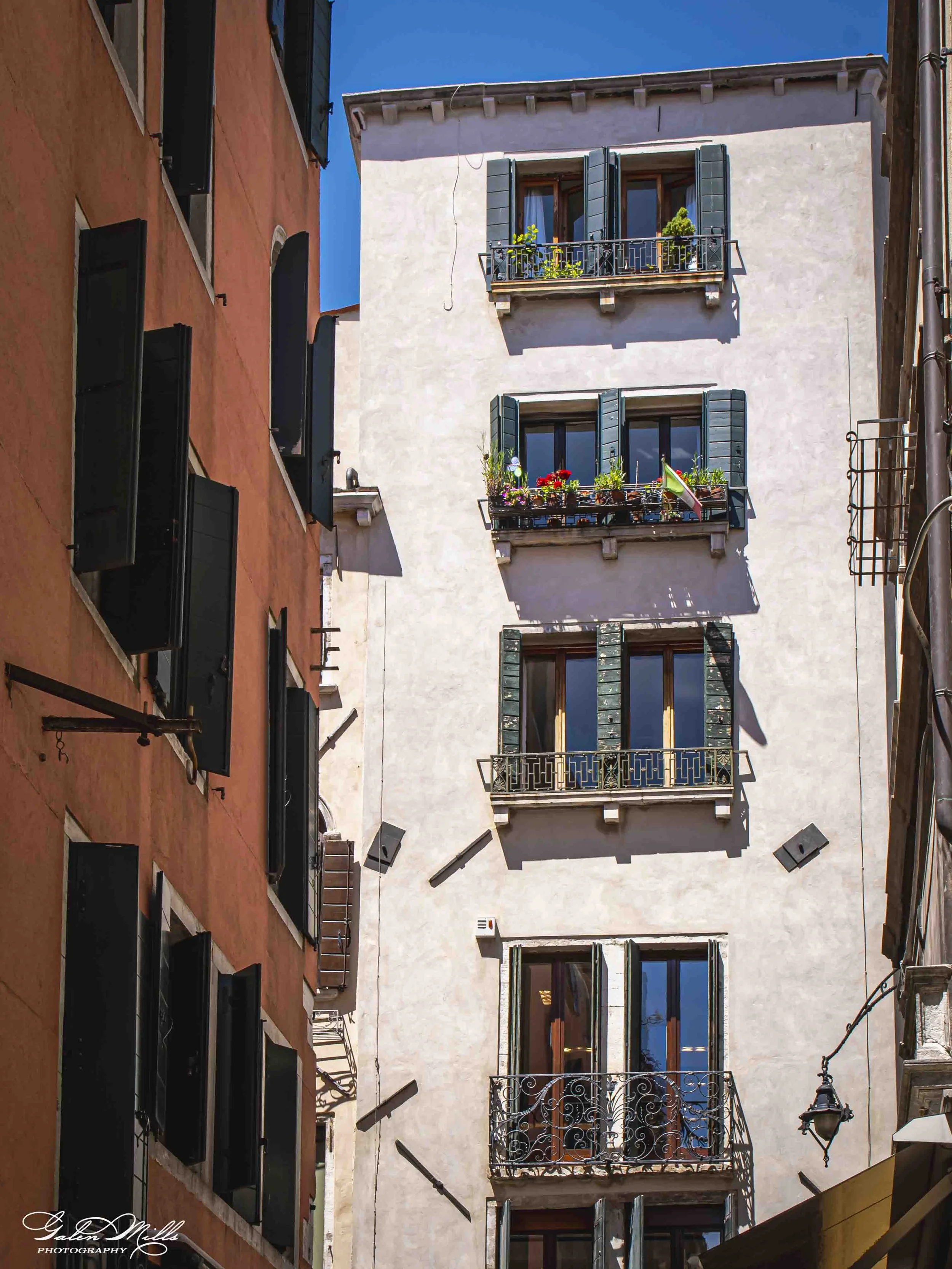 A narrow street viewed from below with colorful buildings on both sides, featuring several balconies with plants and window shutters, and a bright blue sky above.