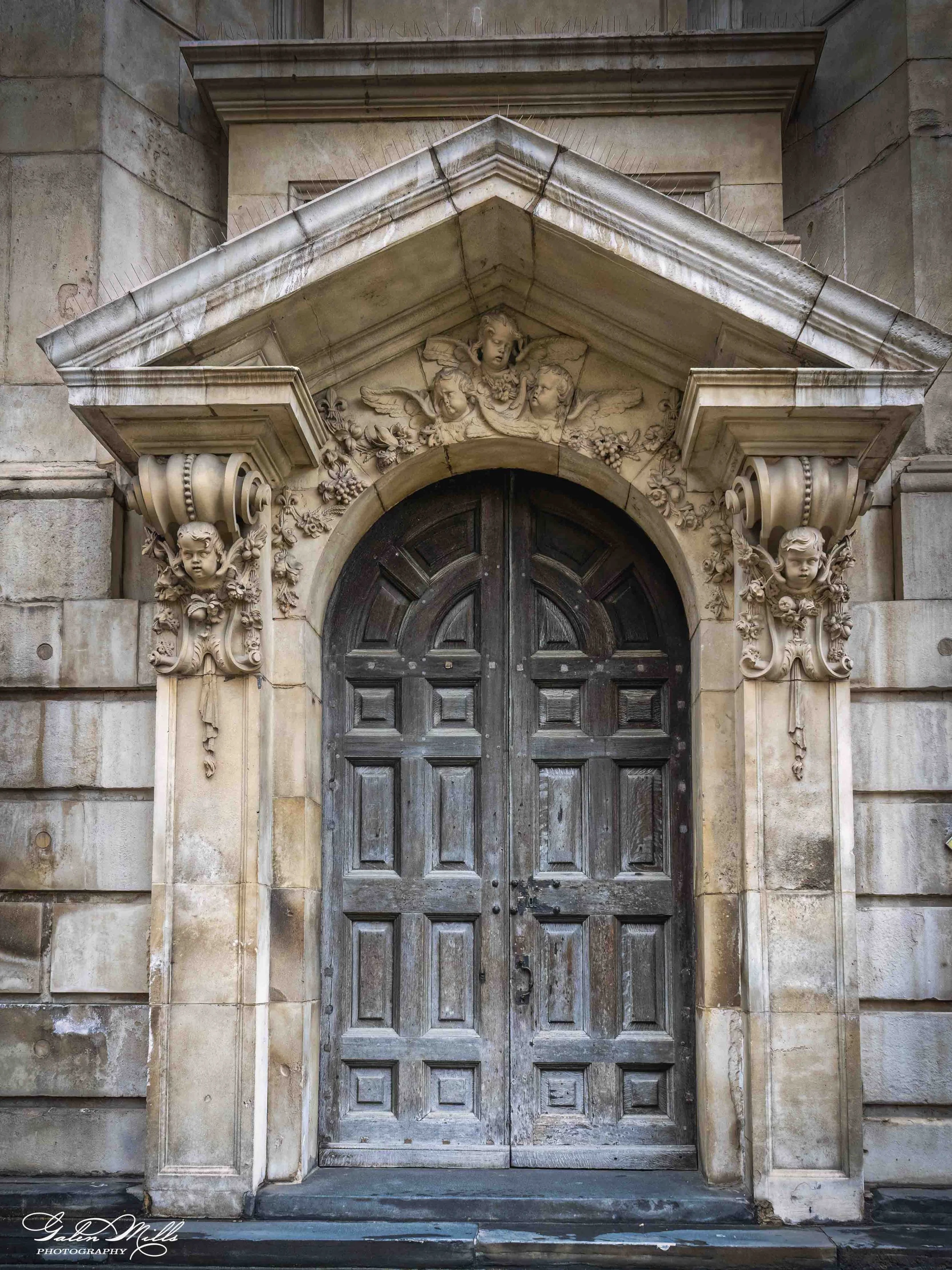 Stone building entrance with ornate carved decoration above and on sides of large wooden double doors. Carvings include cherubs, faces, and floral motifs.