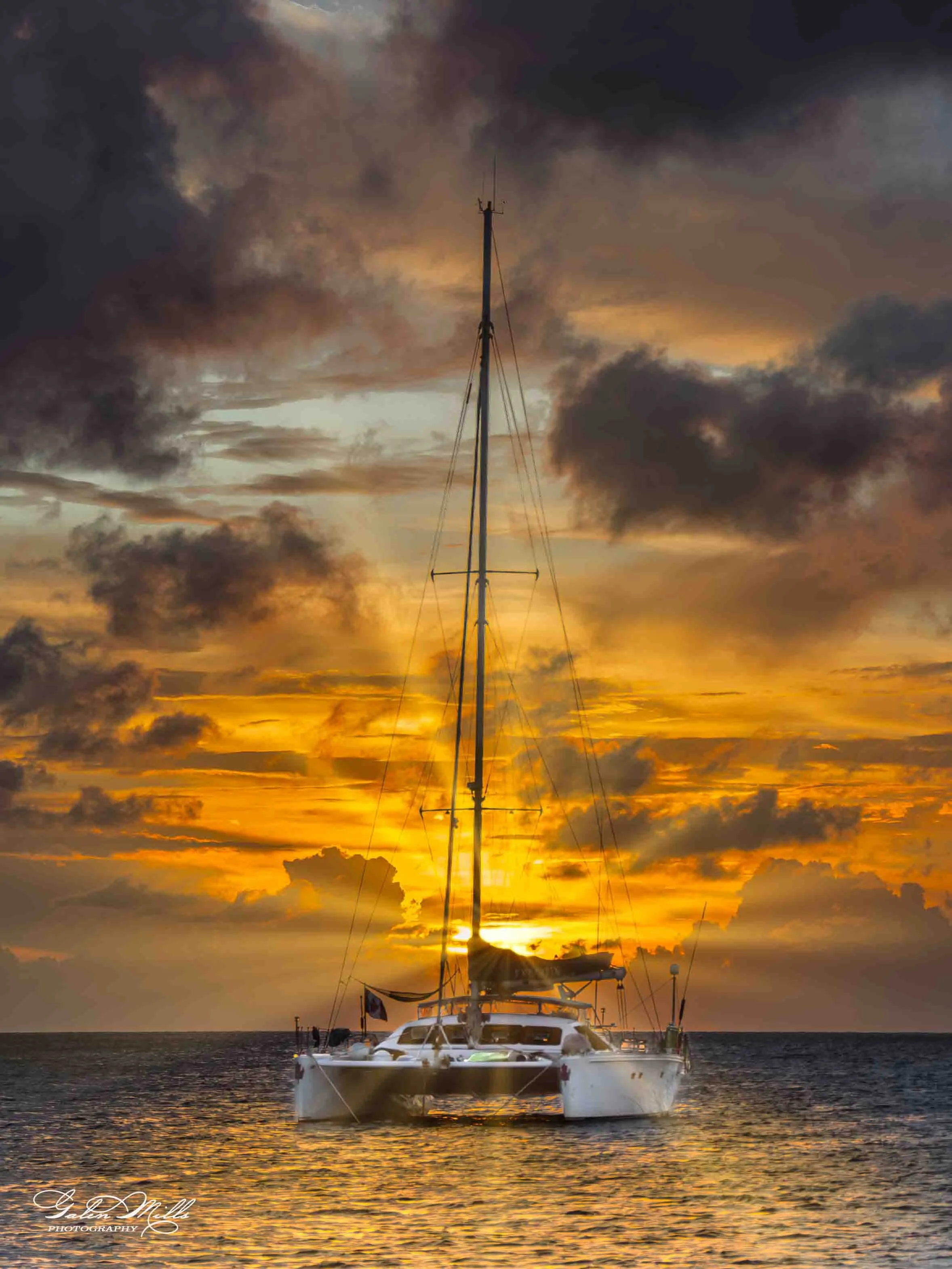 A sailboat on the water during sunset, with a dramatic sky filled with clouds and vibrant orange and yellow colors.