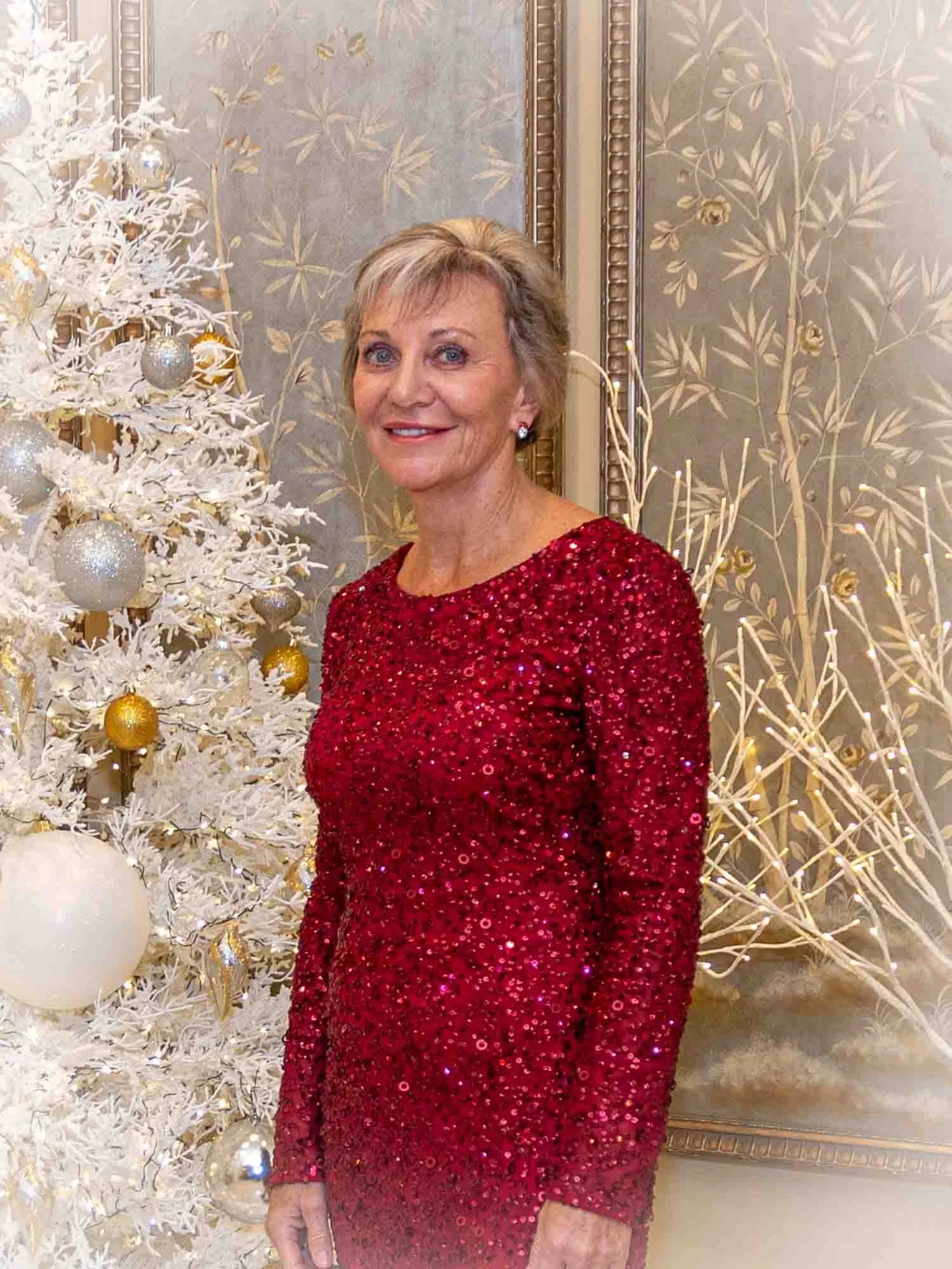 Woman in red sequined dress standing next to a decorated white Christmas tree with silver and gold ornaments in a room with ornate wallpaper.