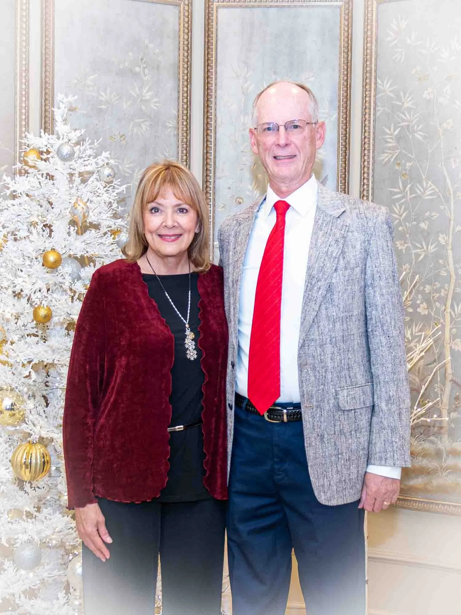 An older couple standing together indoors, smiling in front of a decorated white Christmas tree with gold and silver ornaments. The woman is wearing a red jacket and black outfit, while the man is wearing a gray blazer, white shirt, and red tie.