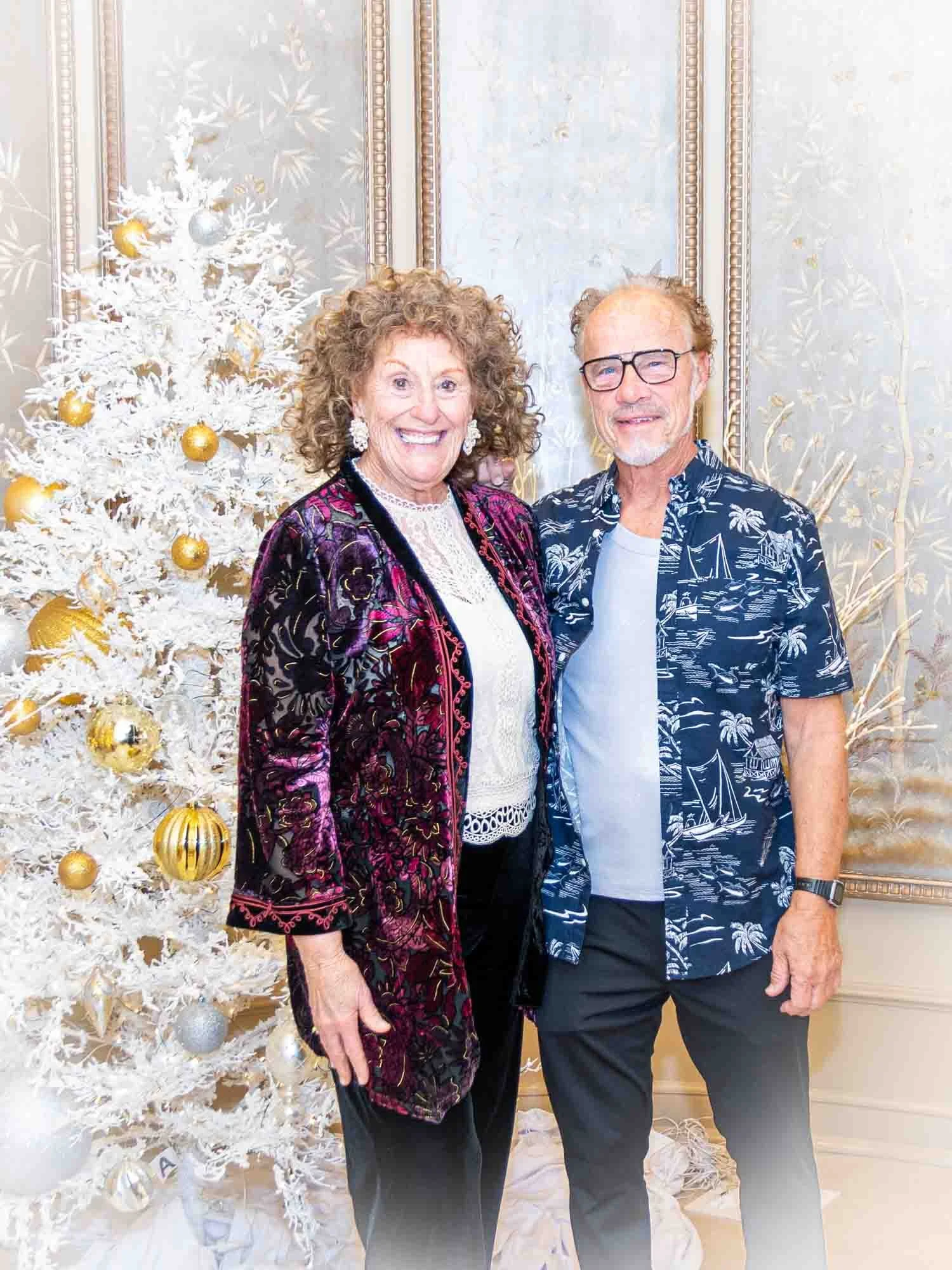 Smiling couple posing in front of a white Christmas tree with gold and silver ornaments. The woman wears a velvet jacket, and the man sports a patterned shirt. Elegant interior setting.