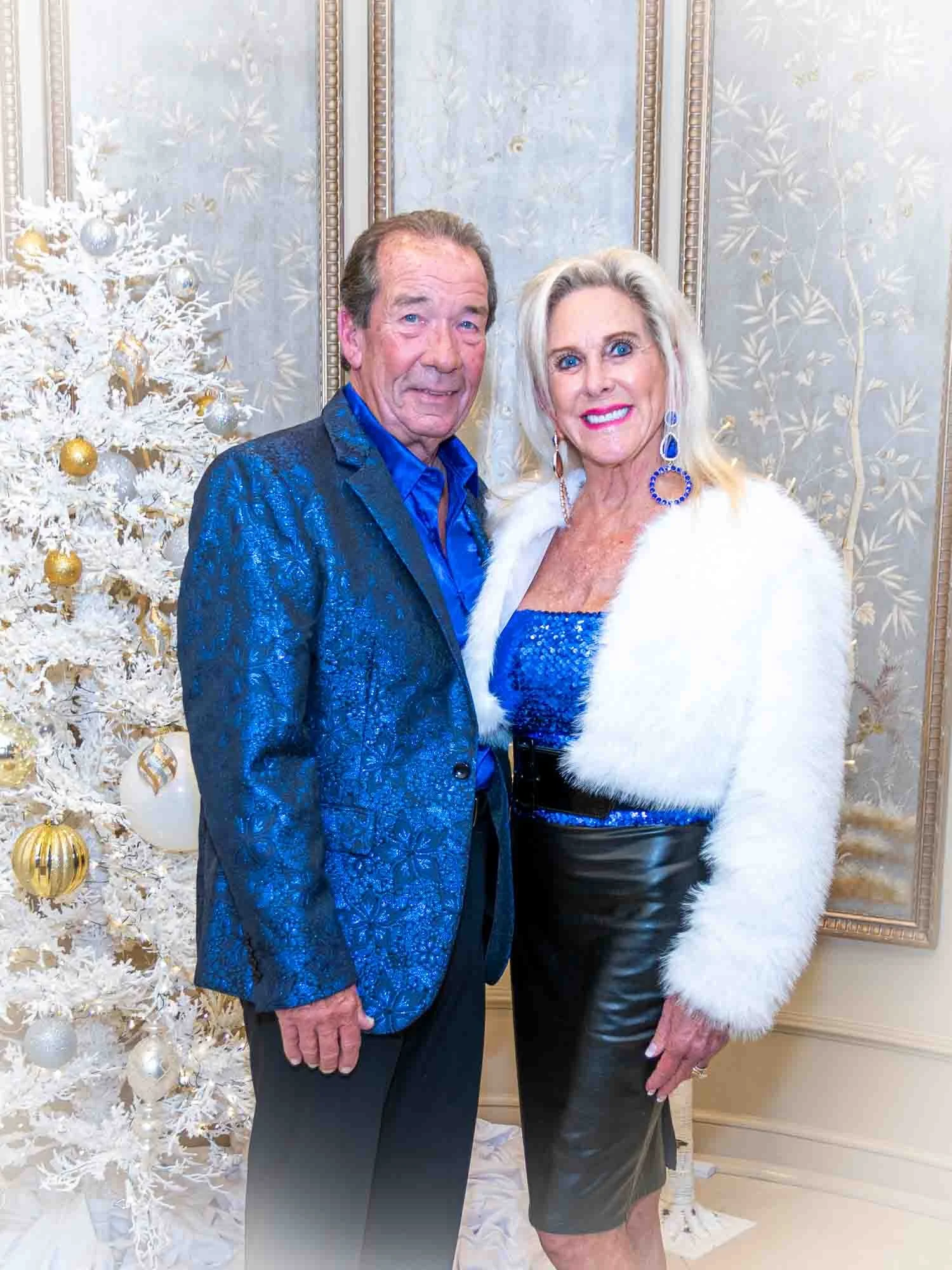 An elegantly dressed couple posing in front of a white Christmas tree with gold ornaments, the man in a blue textured suit and the woman in a white fur coat and blue dress.