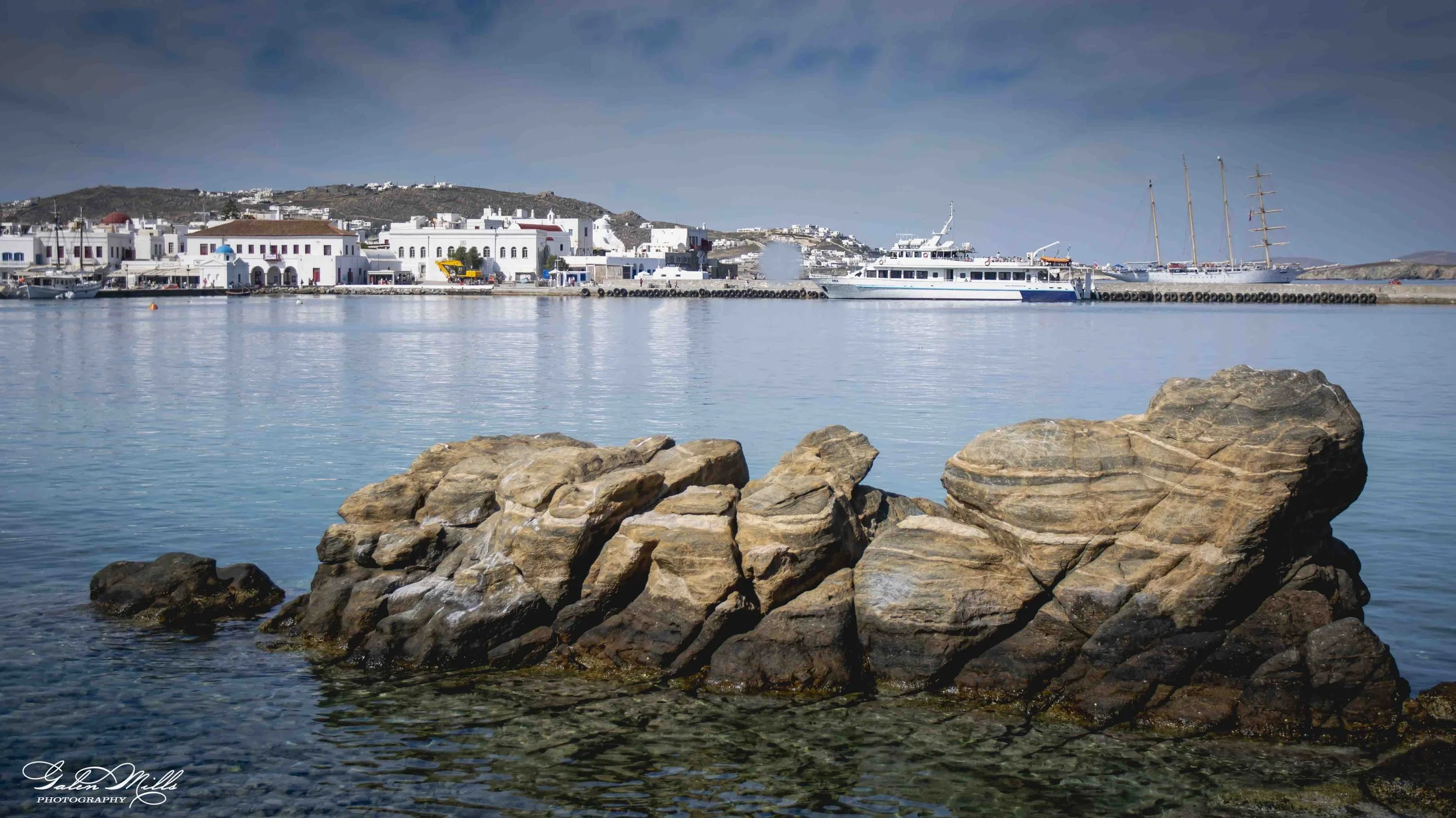Rocks in calm water with white buildings and boats along the harbor, hilly landscape in the background under a cloudy sky.