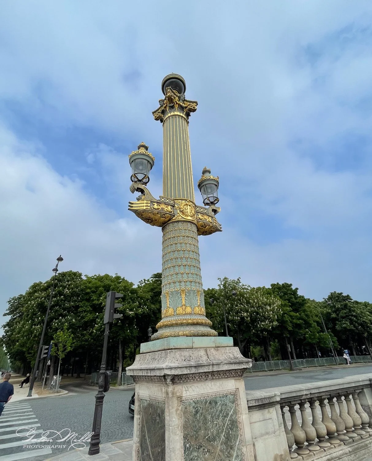 Decorative lamp post with gold embellishments and two lanterns on a stone base, located near a tree-lined street.