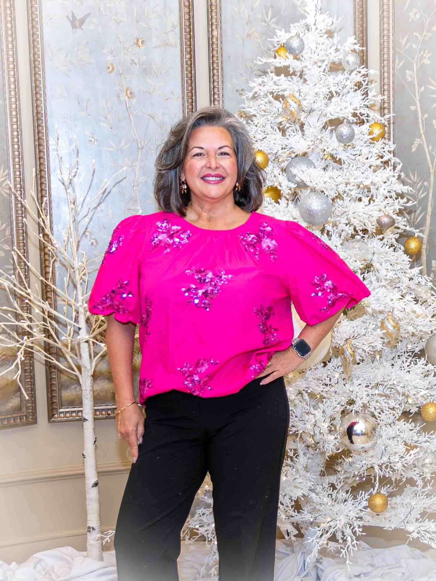 Person in a pink blouse and black pants standing next to a decorated white Christmas tree indoors.
