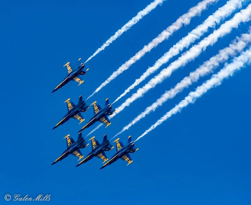 Blue Angels jets in formation flying against a clear blue sky, emitting white smoke trails.
