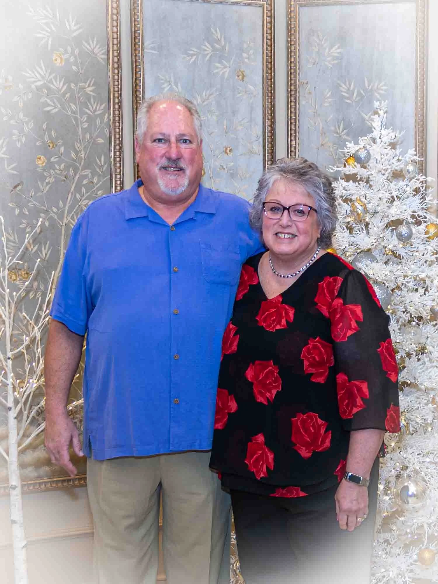 A couple standing in front of a decorative background with a white Christmas tree.