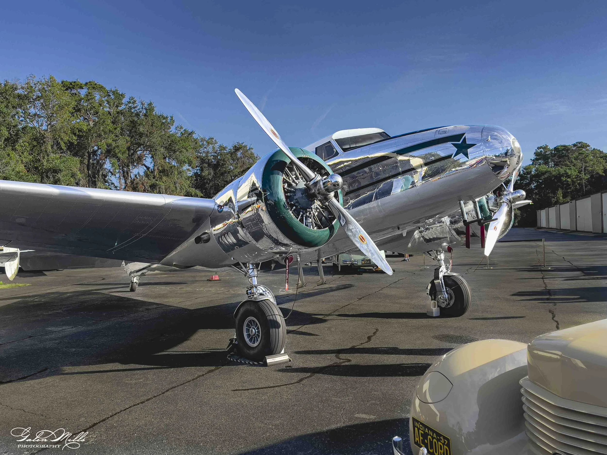 A shiny silver vintage military aircraft with a star insignia on its nose, parked outdoors on a sunny day with a clear blue sky and green trees in the background. The aircraft's propeller is visible, and part of a white car with a visible Indiana lic