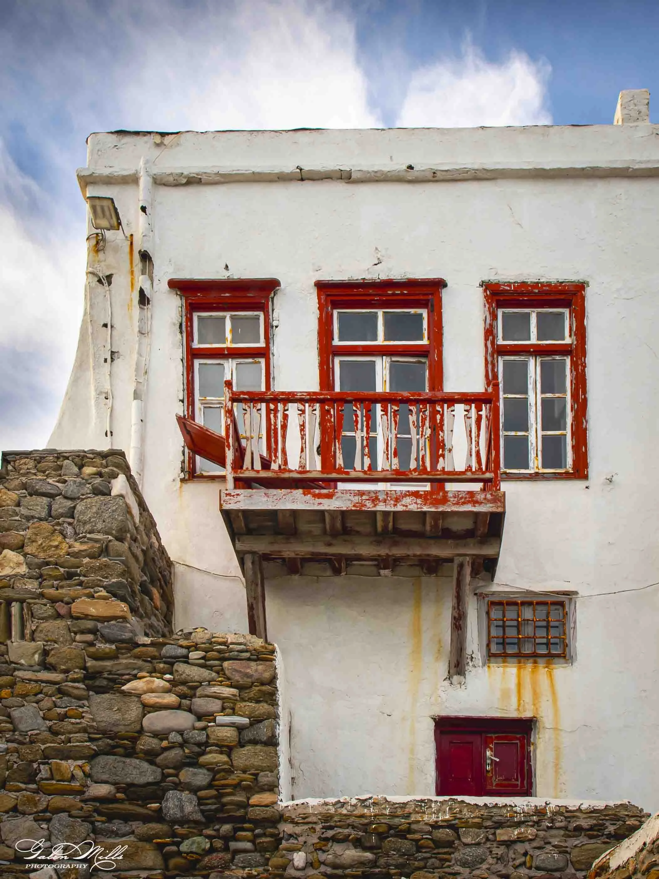 A white building with three red-framed windows and a red door, a small balcony with a red railing, on a stone foundation, against a blue sky with white clouds.
