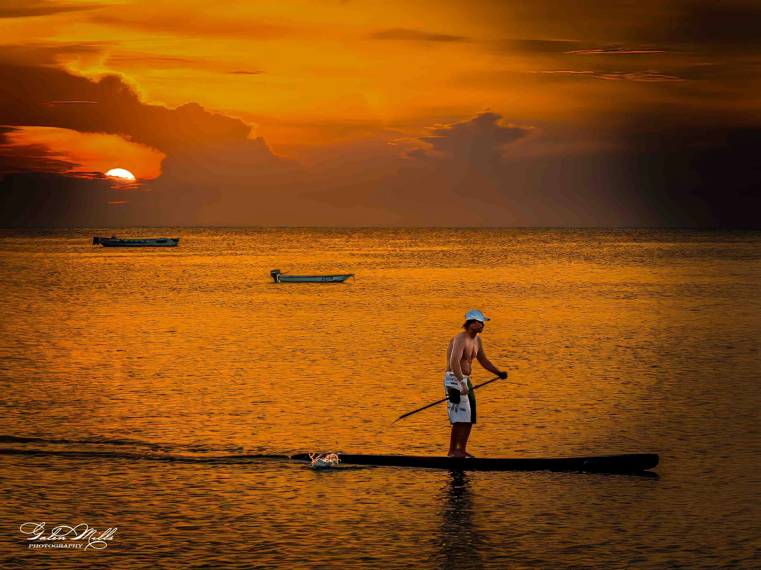A man water surfing on a paddleboard during sunset on the ocean, with two boats floating in the background and a dramatic sky.