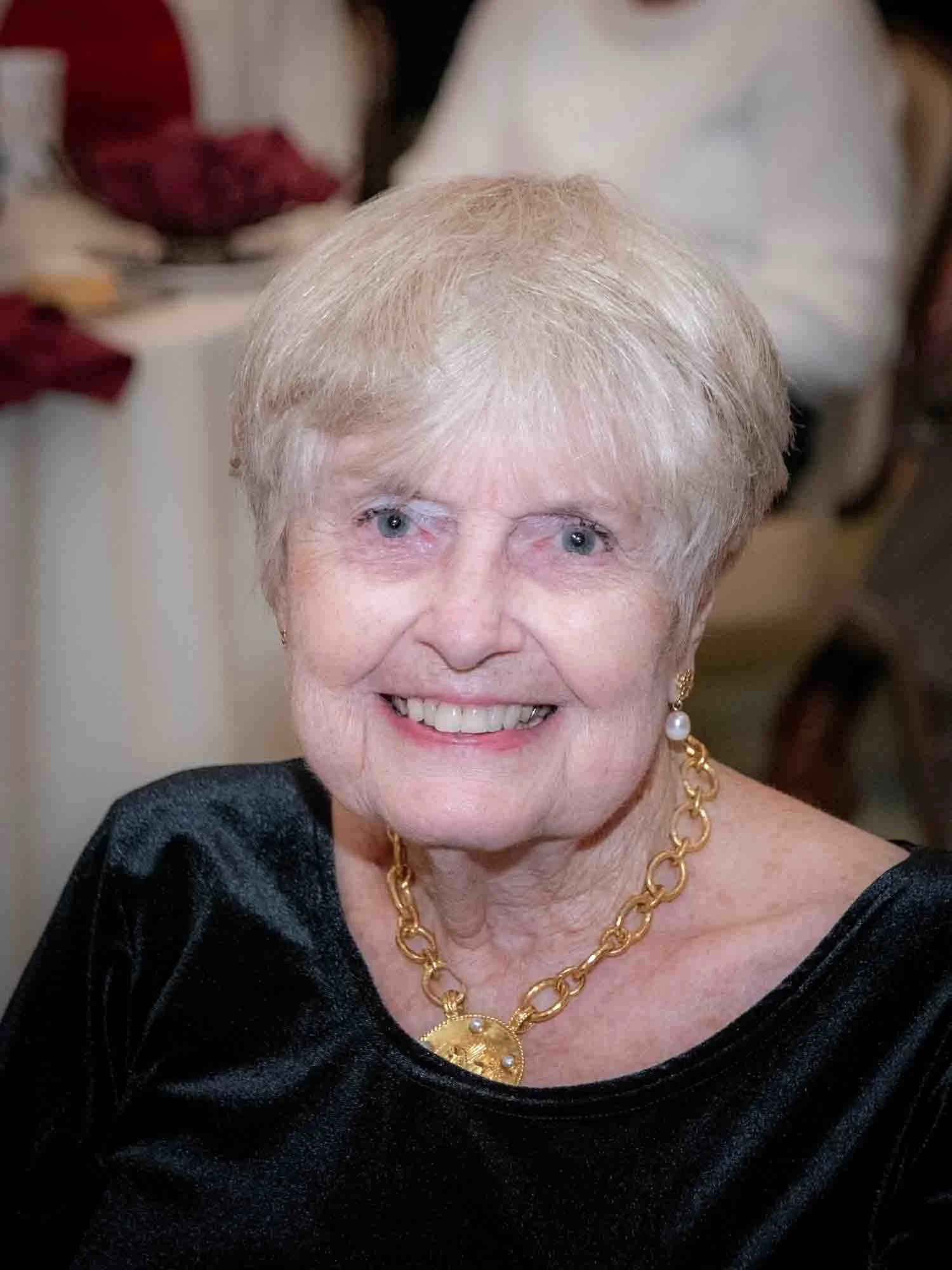 Elderly woman with short hair, wearing a black top and gold necklace, smiling at an event.