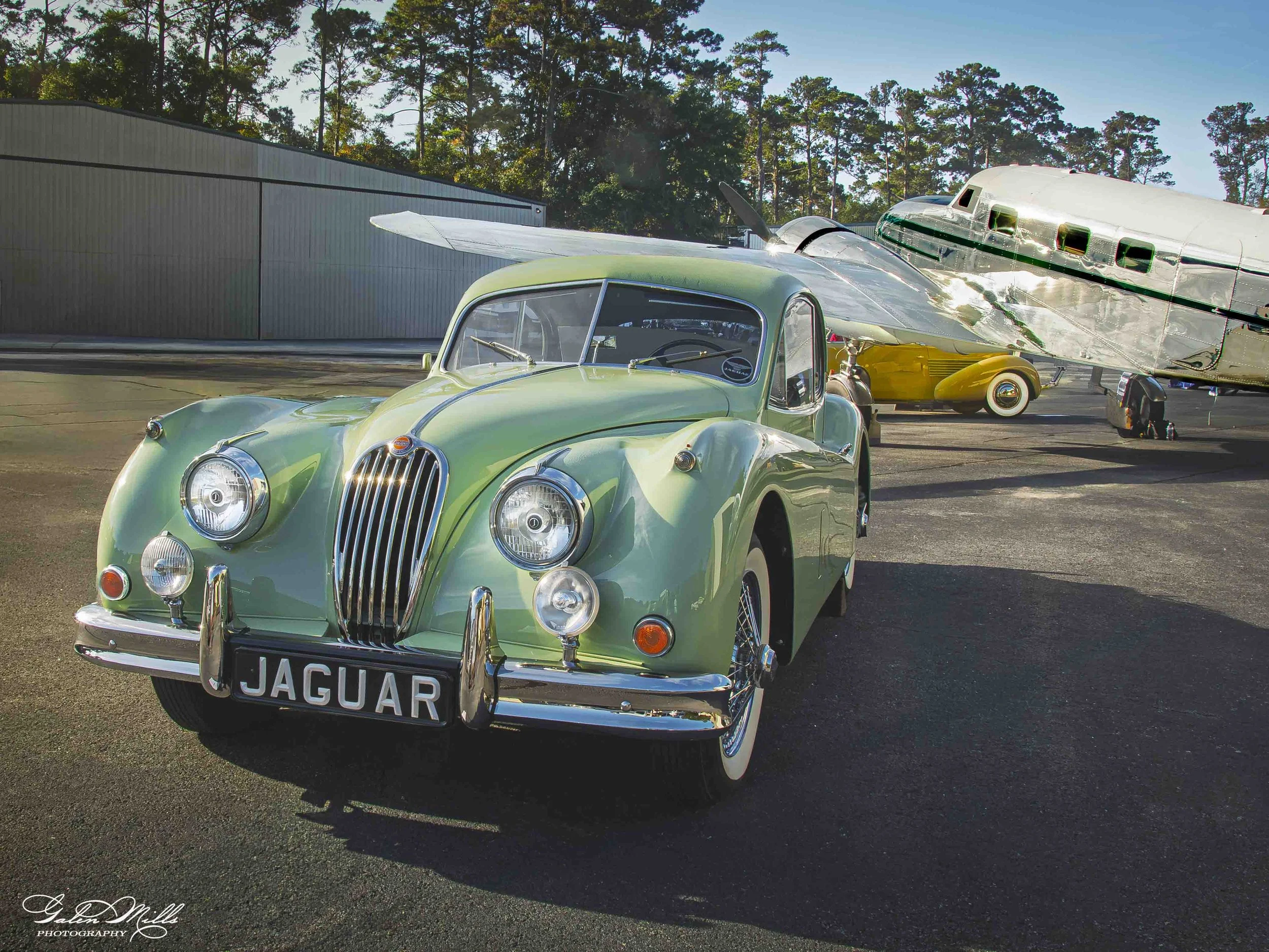 A vintage light green Jaguar XK120 car with a chrome grille, parked outdoors on a paved surface. Behind it, a silver aircraft with green details is parked. The background features tall trees and a clear sky.