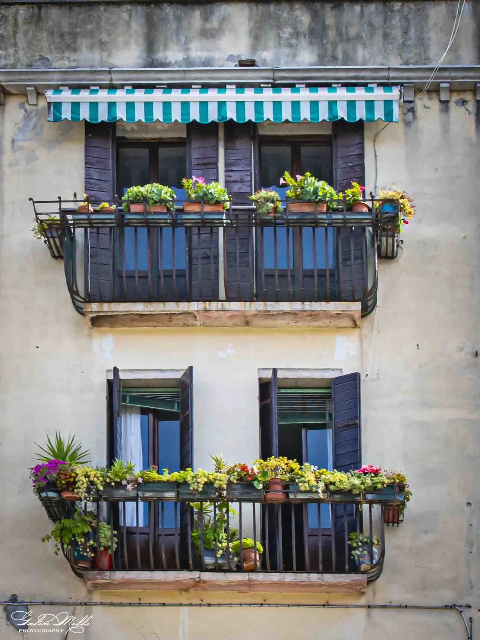 Two balconies with flower boxes on a cream-colored wall, one above the other, decorated with various colorful flowers and green plants.