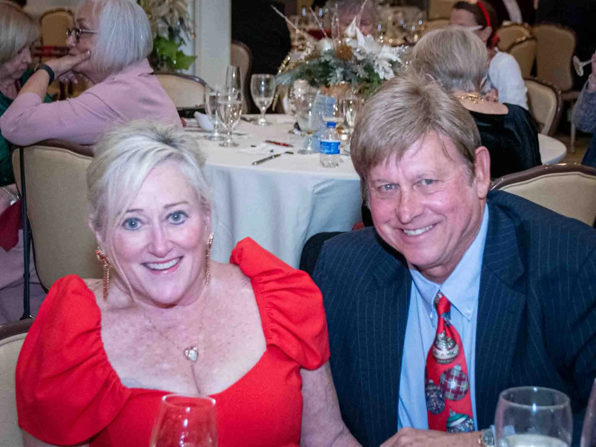 A smiling couple at a formal event, with the woman in a red dress and the man in a suit with a festive tie. They are seated at a table in a banquet setting with other guests in the background.