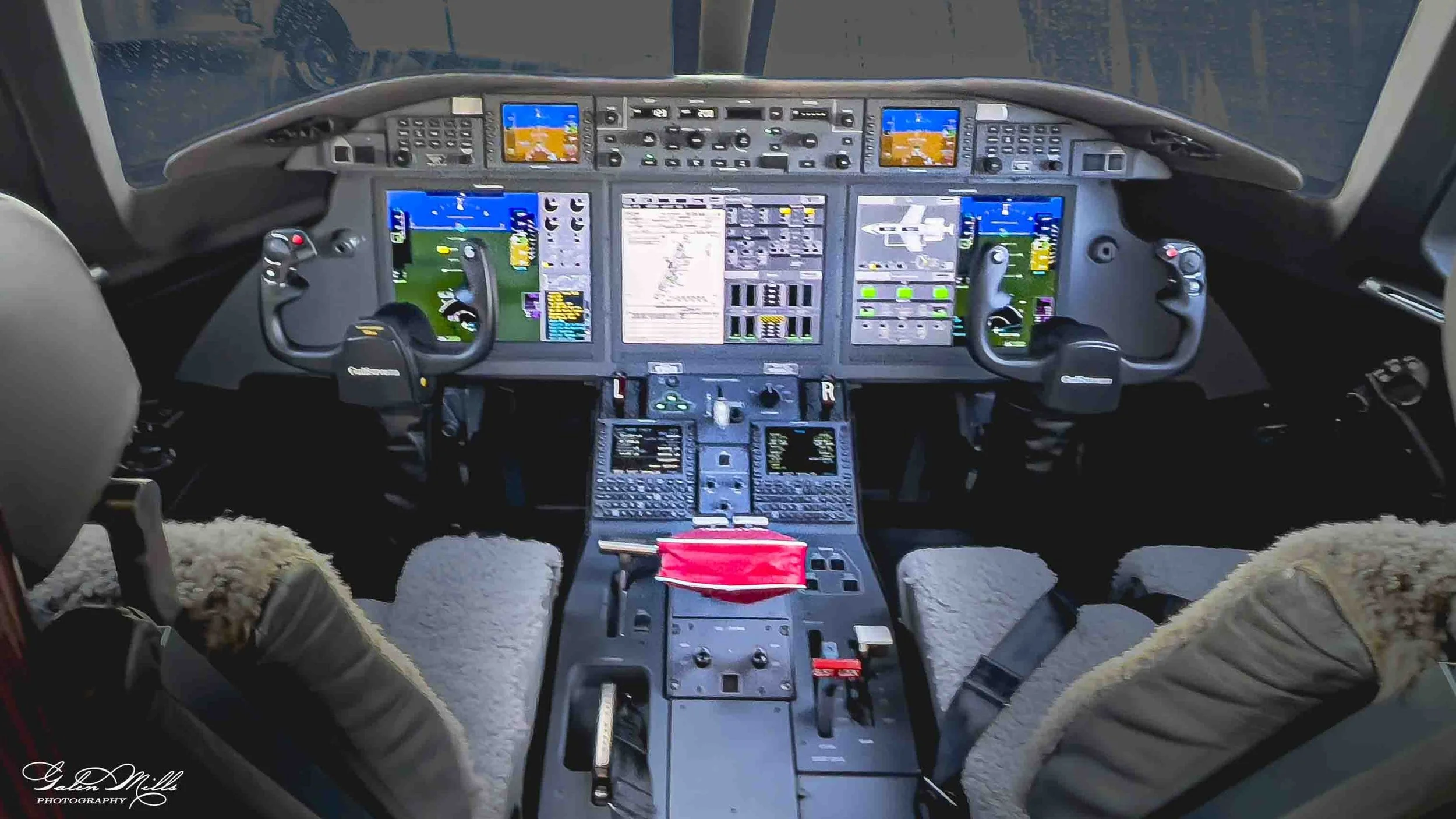 View of an airplane cockpit with multiple digital screens, controls, and two pilots wearing shearling-lined jackets, seated in front of the controls.