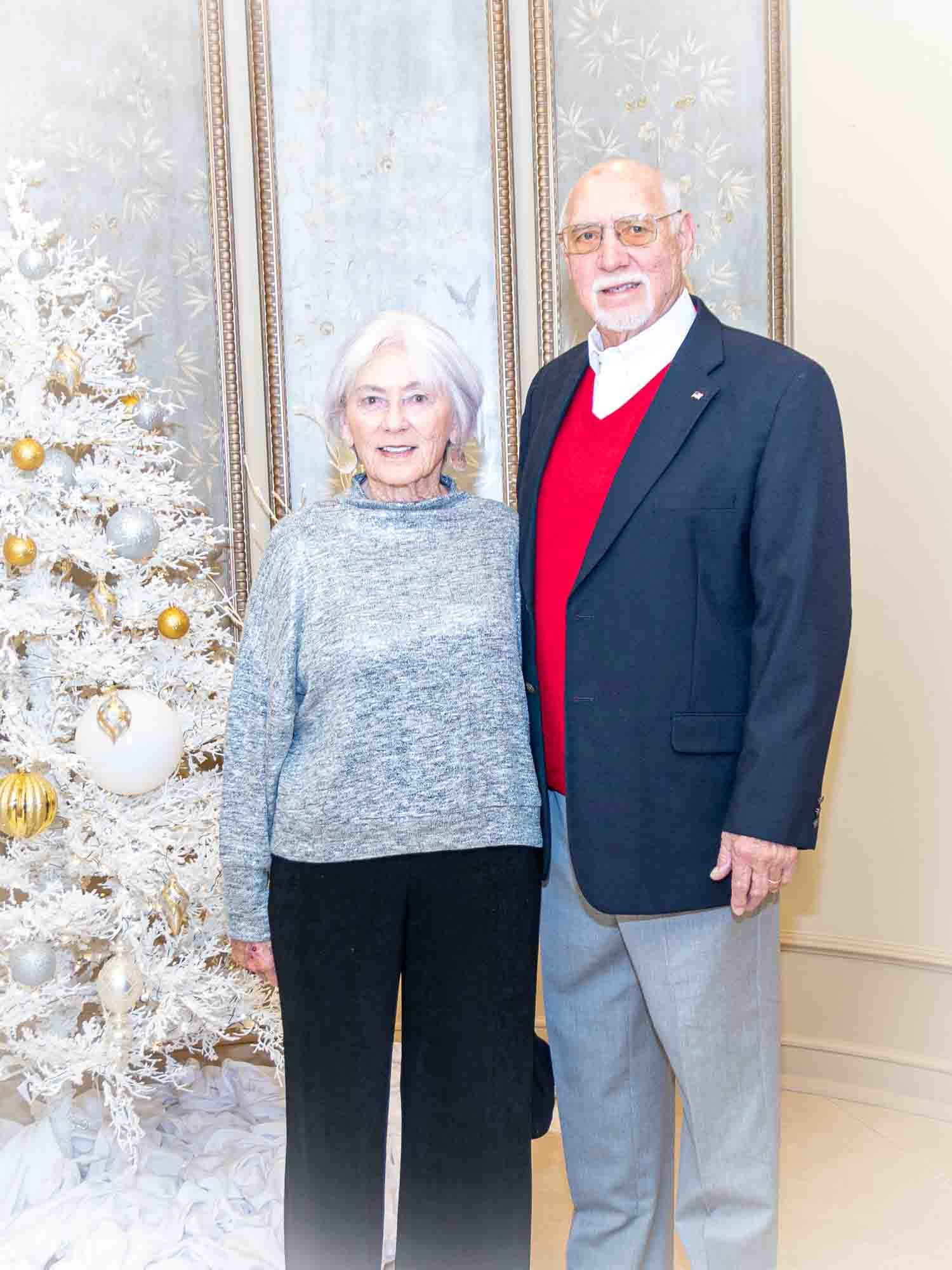 Elderly couple standing in front of a decorated white Christmas tree indoors, woman wearing a gray sweater and black pants, man wearing a red sweater and dark blazer.