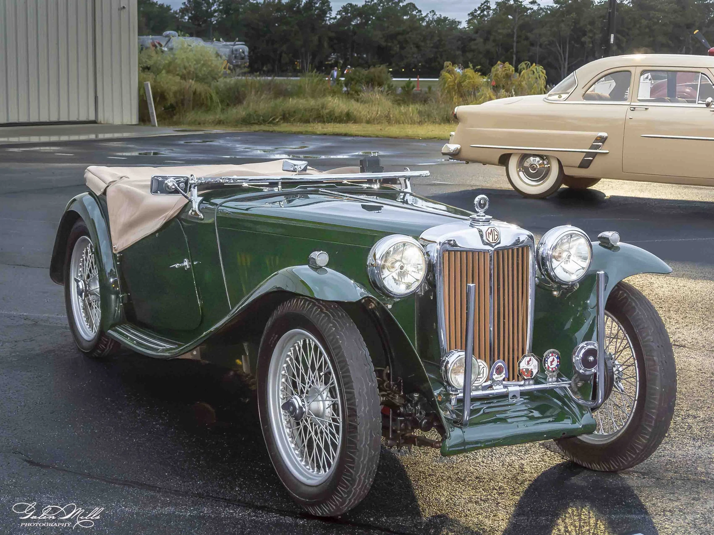 A classic green MG car with a beige cover on the rear, parked beside a beige vintage car. The setting appears to be an outdoor area with trees and a building in the background.