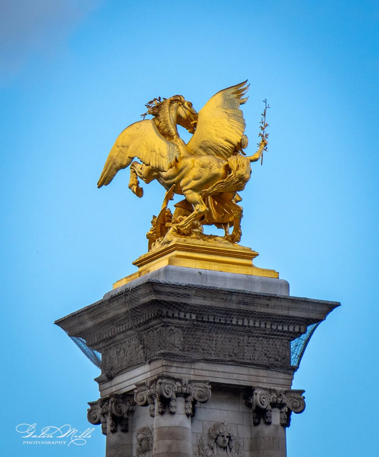 Golden statue of a Pegasus on a stone pedestal with ornate carvings against a blue sky.
