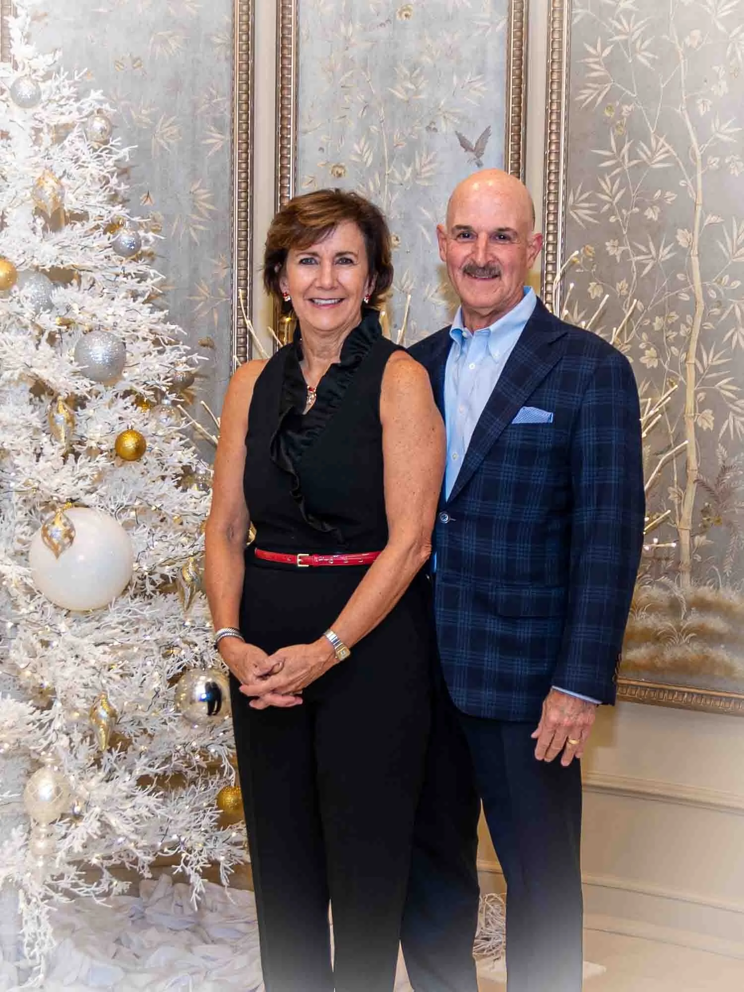 A man and a woman smiling and standing in front of a white Christmas tree decorated with gold and silver ornaments, in a room with ornate wallpaper.
