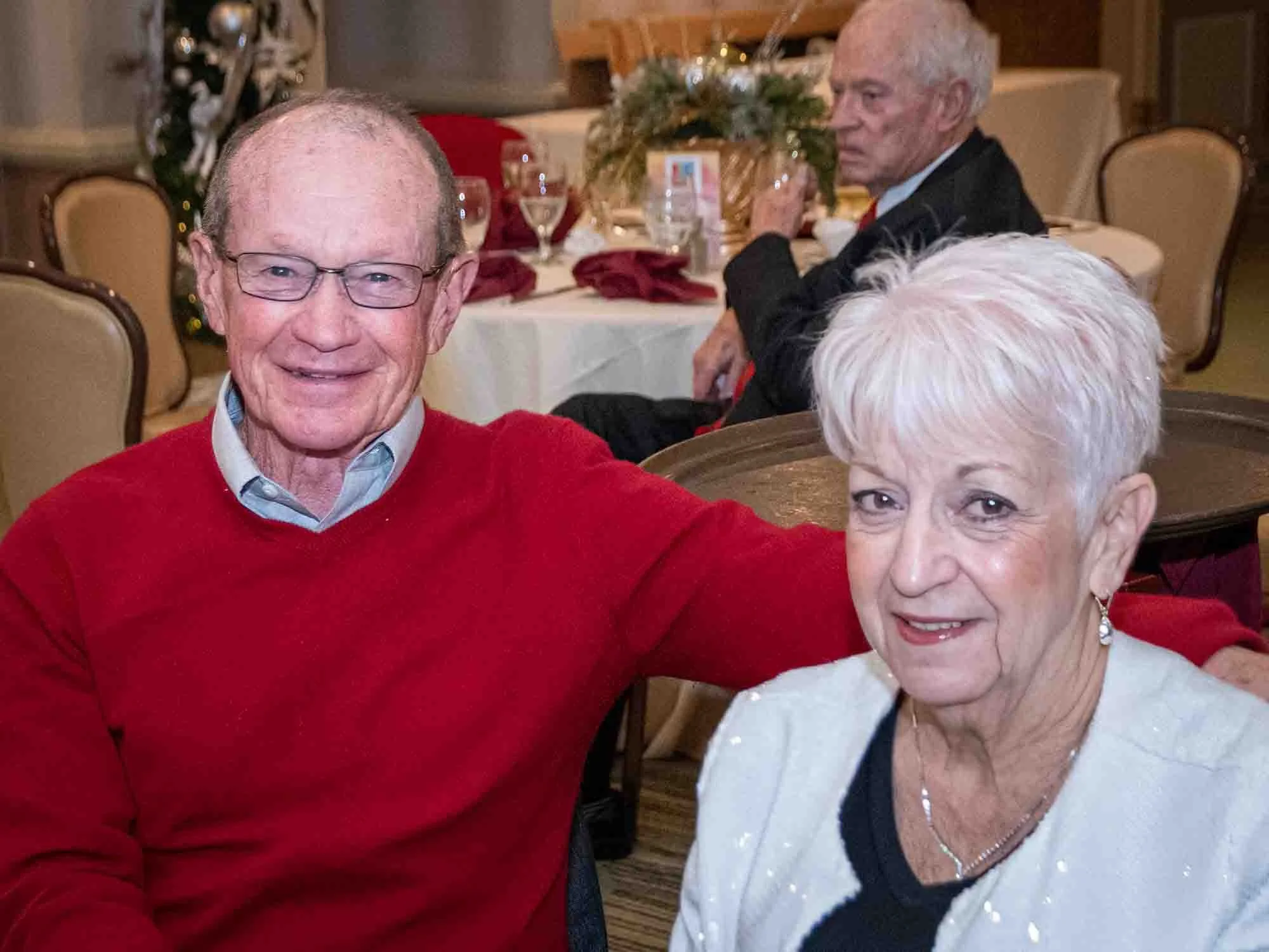 An older man and woman are posing for a photo in a banquet setting, with tables set for a meal in the background. The man is wearing a red sweater and glasses, while the woman has short white hair and is wearing a light-colored jacket. Another elderl