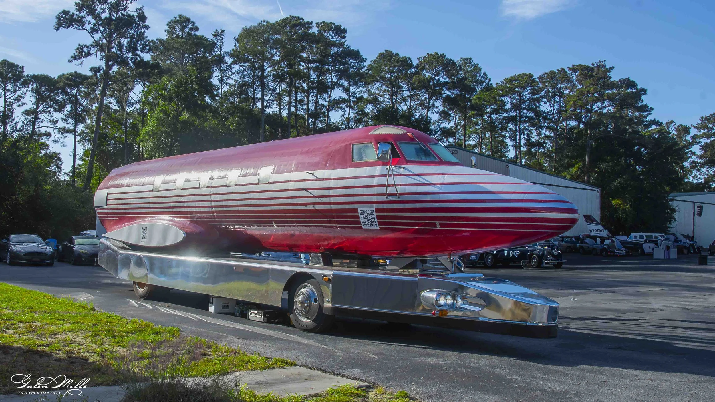 A red Elvis Airplane with white stripes on a shiny trailer, parked in a lot with cars and surrounded by trees and blue sky.
