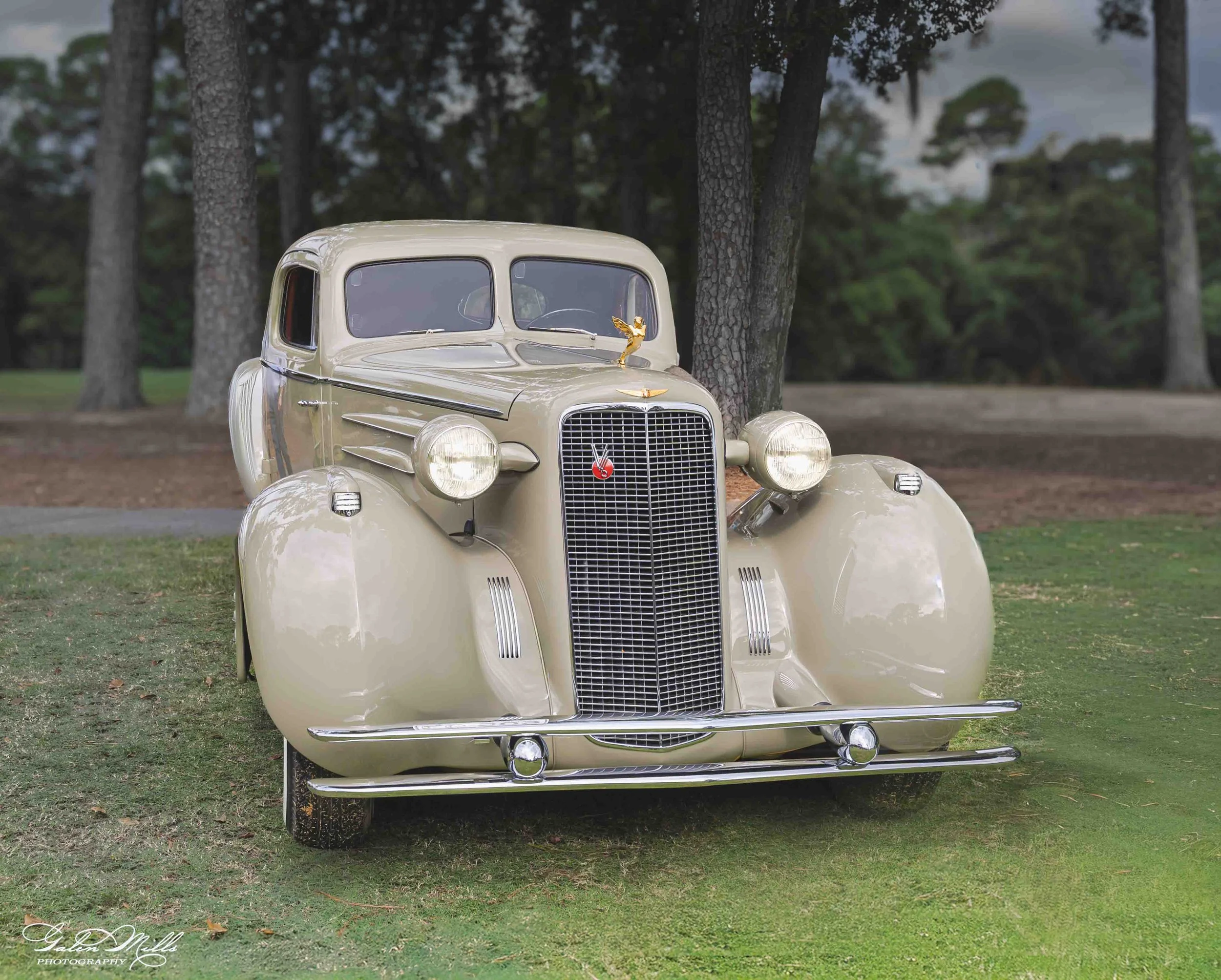 A vintage cream-colored car with decorative gold and black accents on its hood ornament and grille, parked on grass with trees in the background.