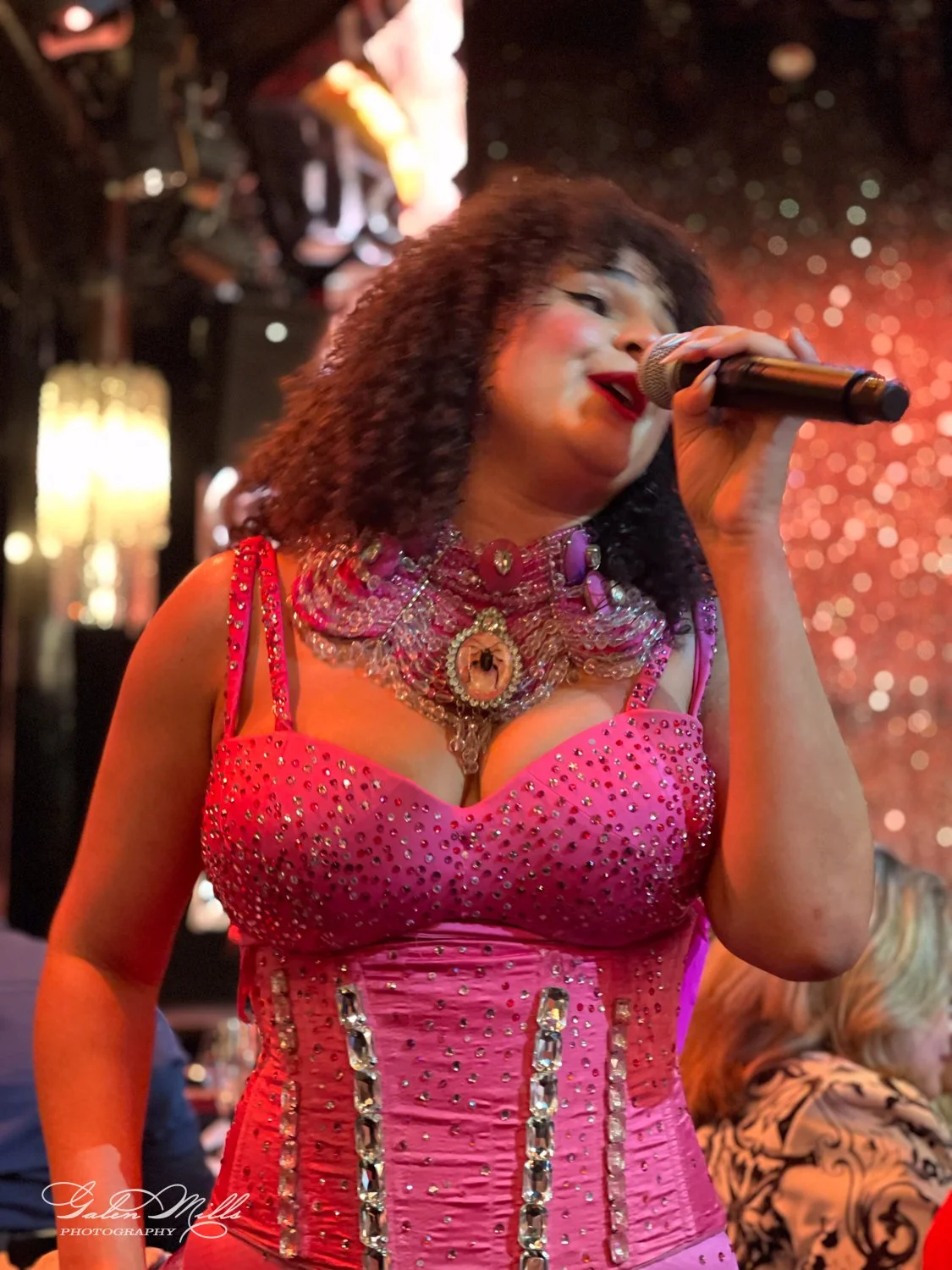 A performer singing into a microphone wearing a sequined pink dress with ornate jewelry on stage, with a sparkling background.