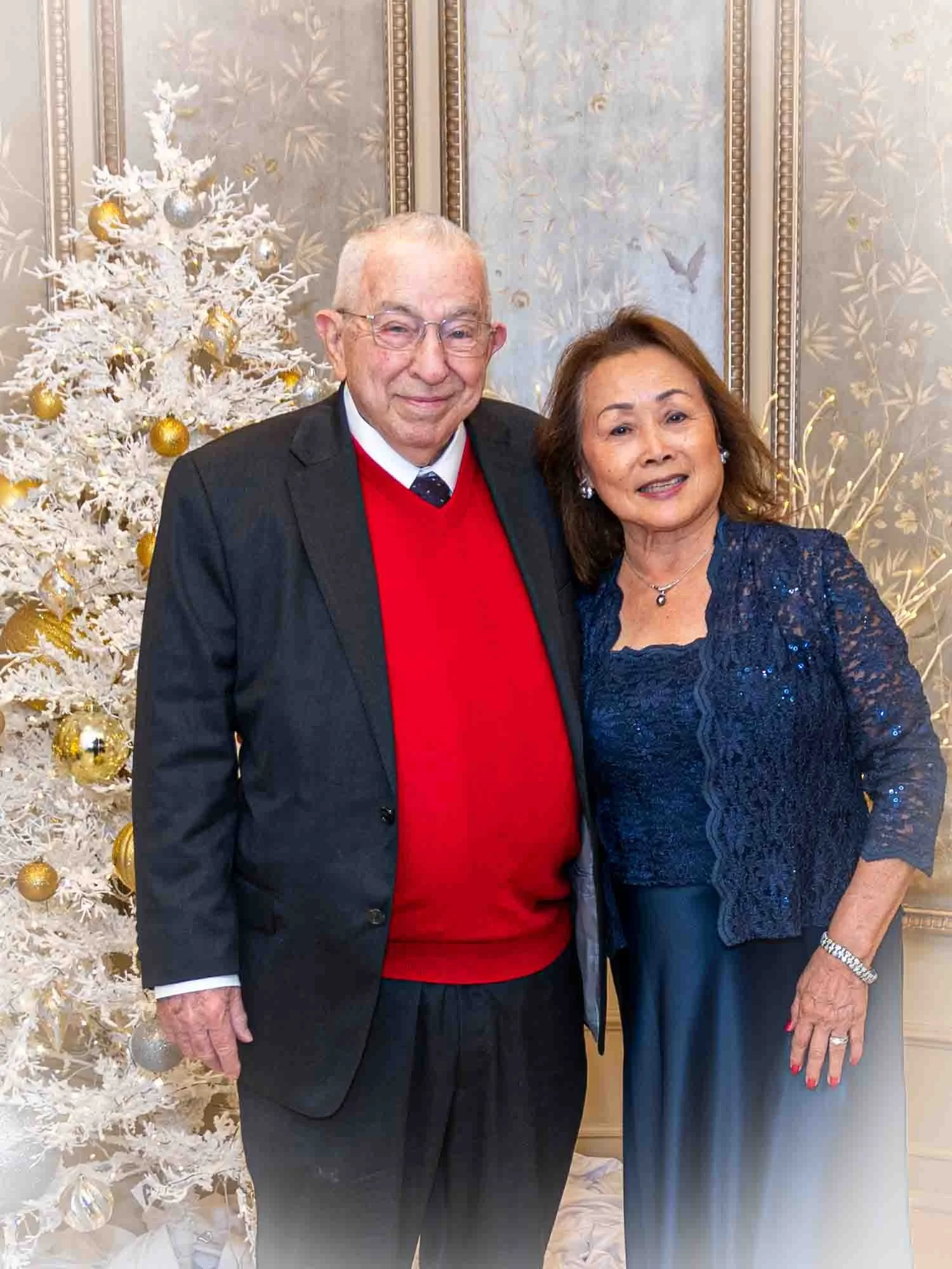 An elderly couple posing next to a white Christmas tree with gold and silver ornaments. The man is wearing a red sweater under a black suit, and the woman is in a navy dress and jacket. The background features elegant, patterned wallpaper.
