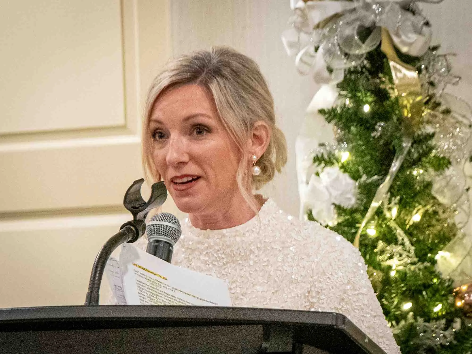Woman speaking at a podium with a microphone, holding papers, in front of a decorated Christmas tree.