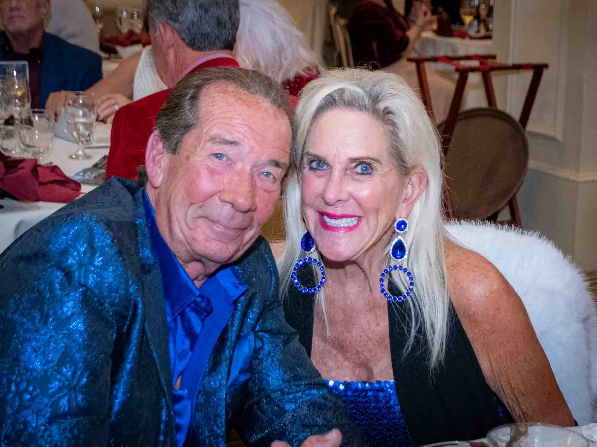 Smiling elderly couple at a formal event, wearing blue and black outfits, surrounded by dining tables.