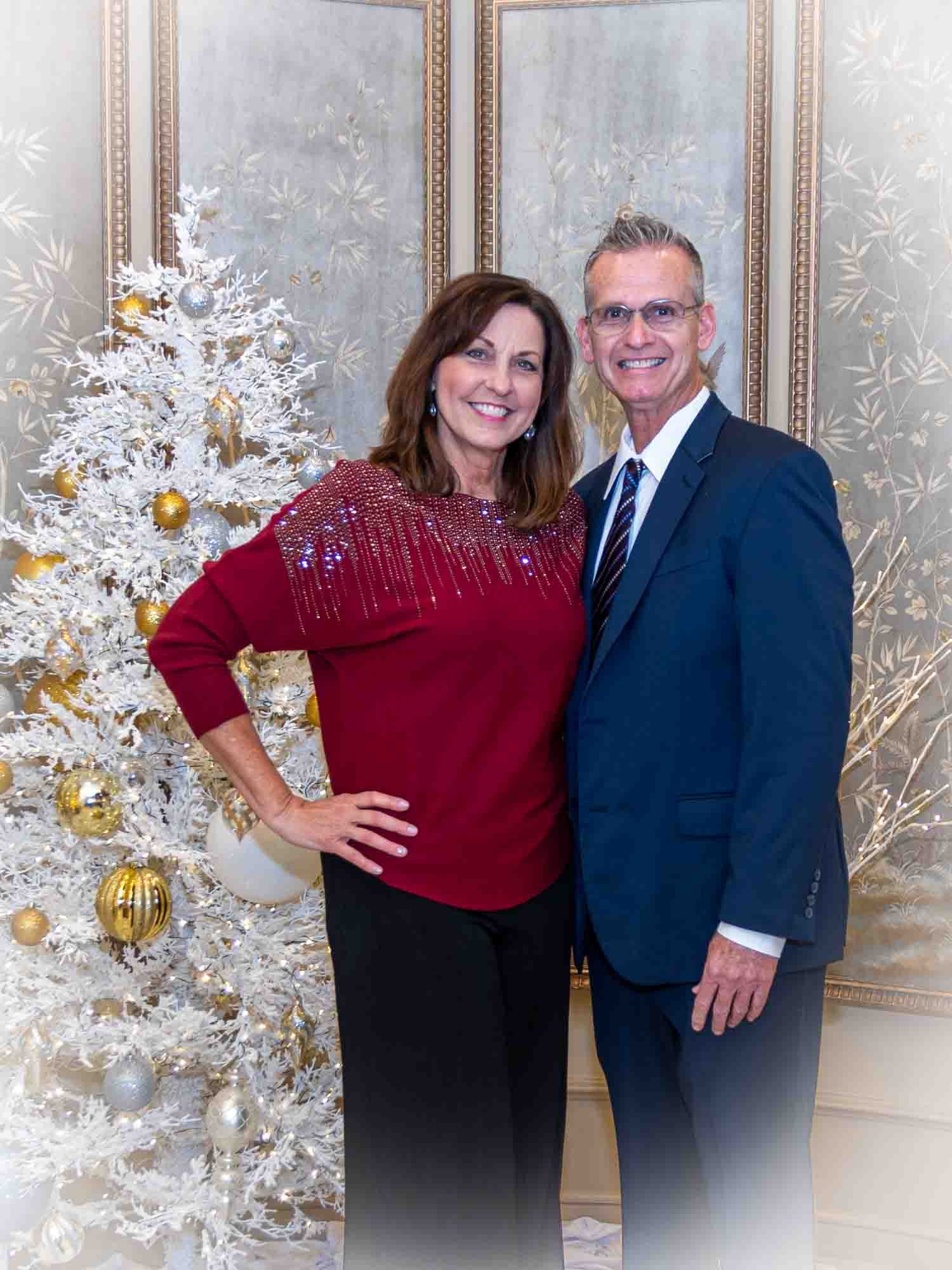 A man and woman posing in front of a white Christmas tree with gold and silver ornaments. The woman is wearing a red blouse and black pants, and the man is in a blue suit with a tie. The background features decorative wall panels.