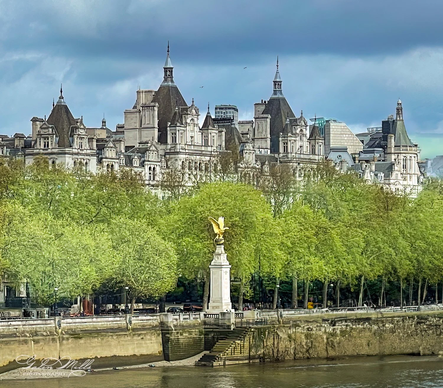Historical buildings with ornate architecture behind lush green trees, a golden eagle statue on a pedestal near a riverbank, under a cloudy sky.