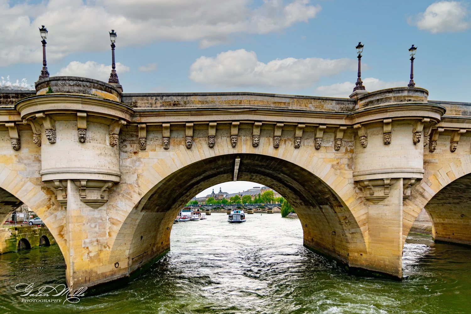 Stone bridge with decorative carvings and arches over a river, boats passing underneath, and buildings in the background.