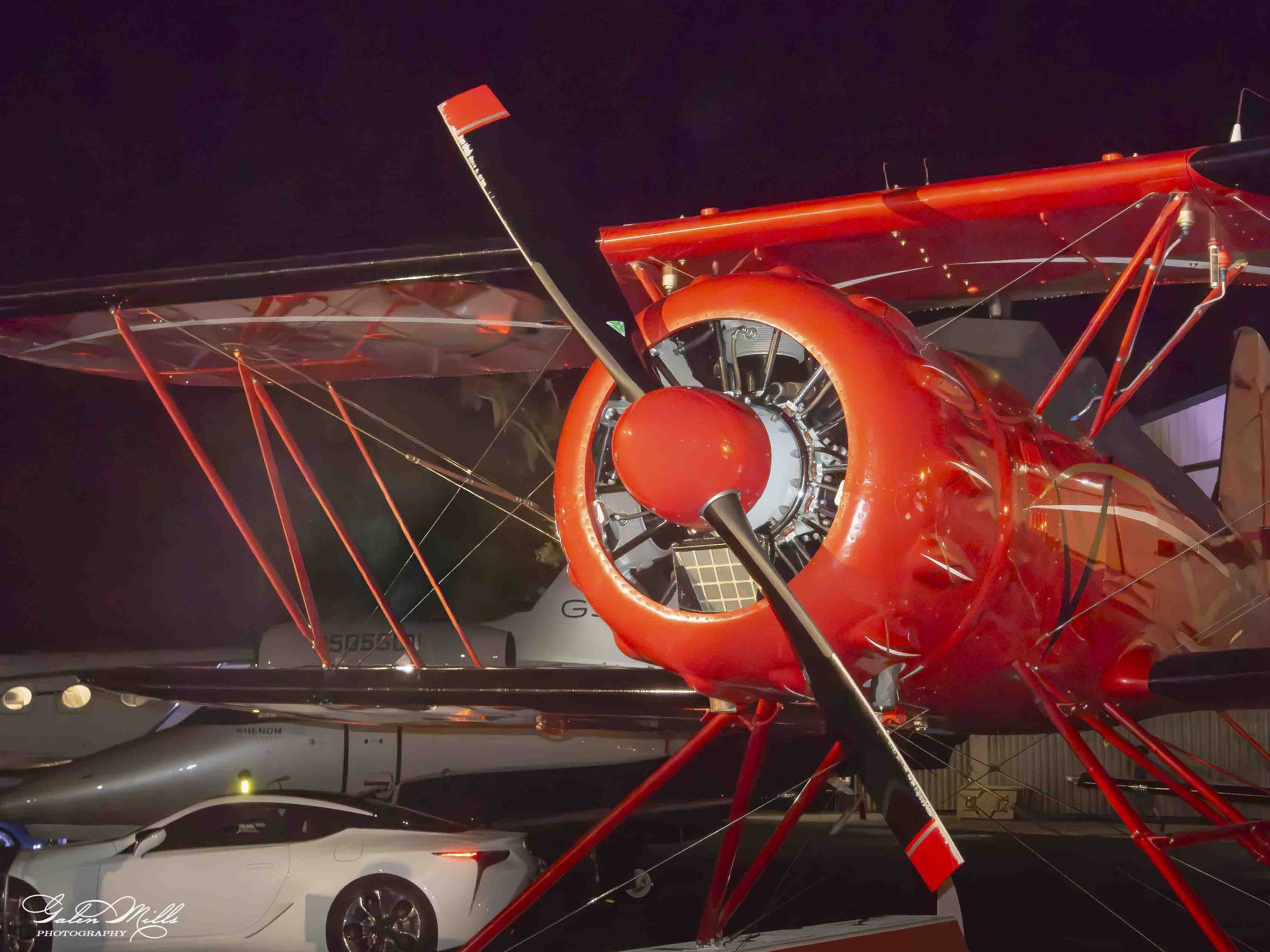 A red aircraft with exposed framework and a large circular engine at the front, parked indoors at night, with other vehicles and objects in the background.