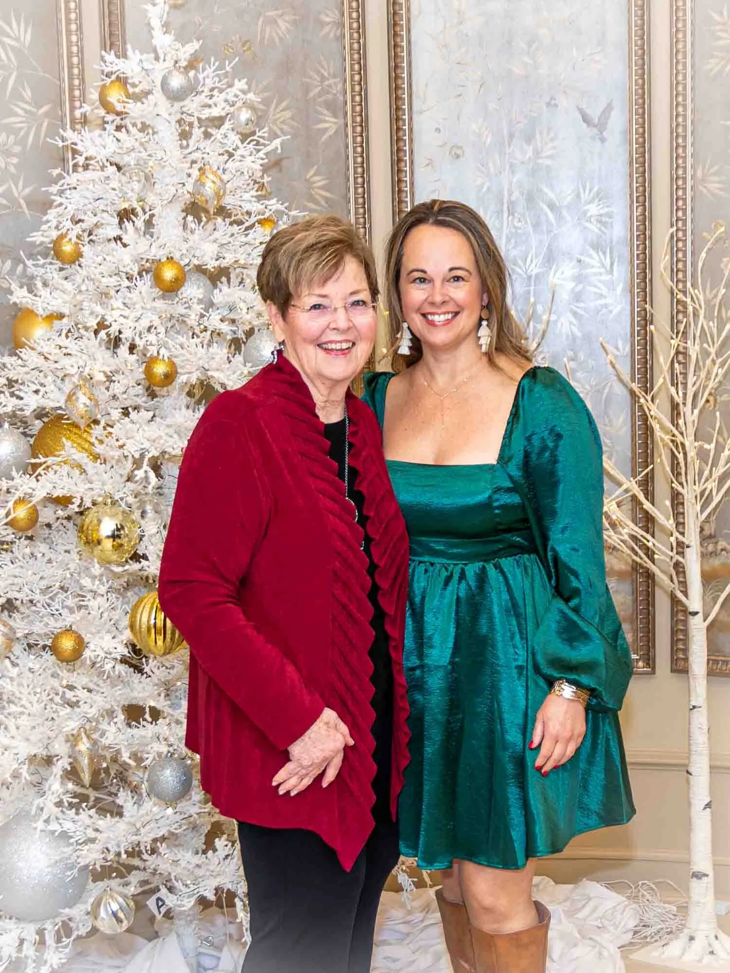 Two women smiling in front of a white Christmas tree decorated with gold and silver ornaments. One woman is wearing a red jacket, and the other is wearing a green dress and brown boots. They are posed in a festive indoor setting.
