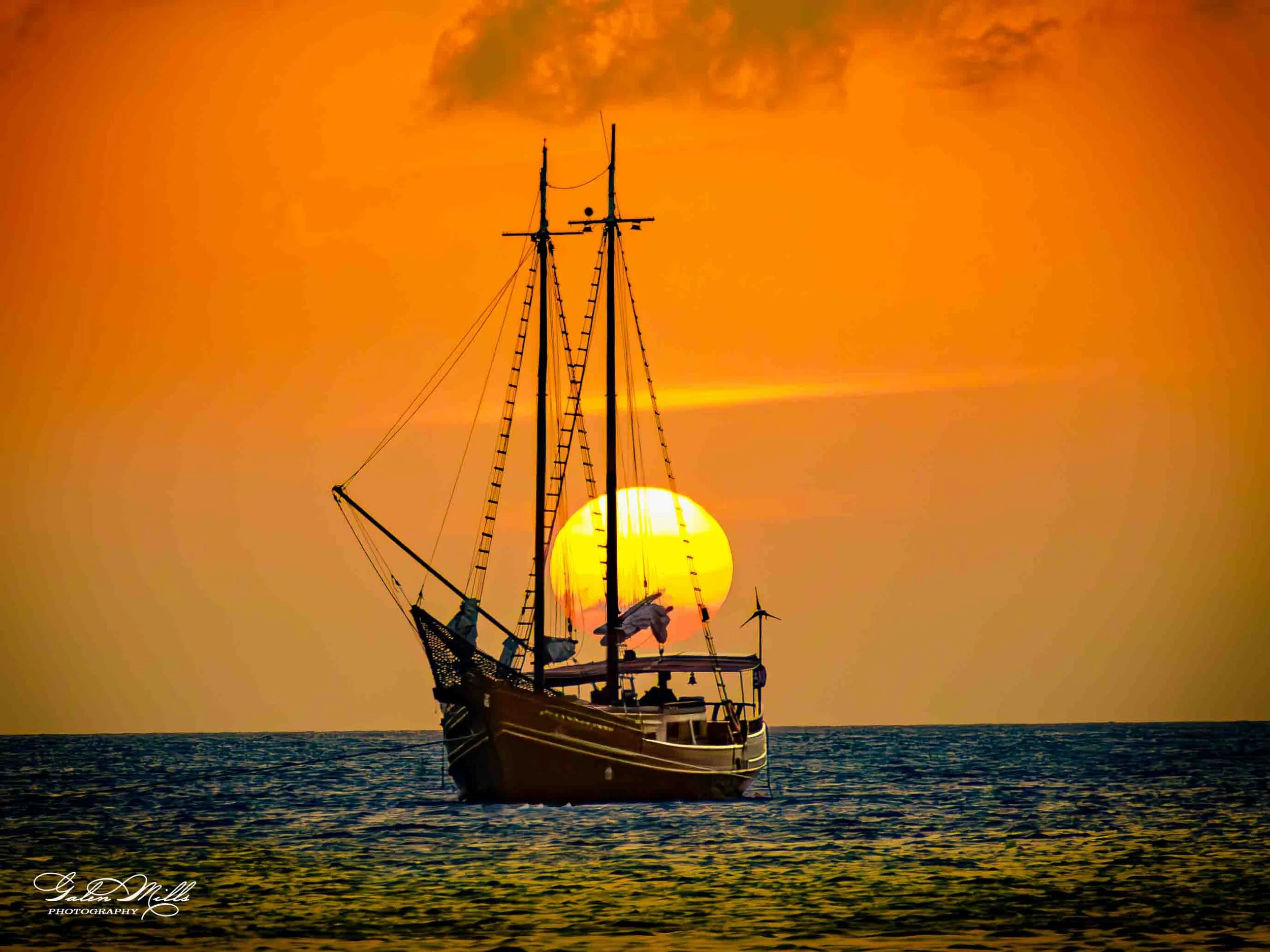 A sailing ship on the ocean during sunset with an orange sky and large setting sun.
