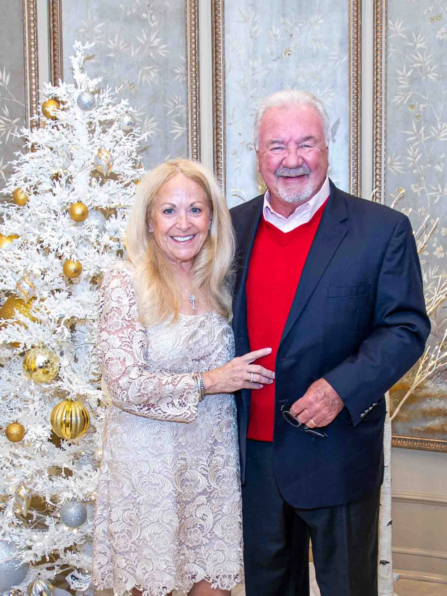 Elderly couple standing in front of a white and gold decorated Christmas tree, smiling and dressed formally; the woman in a lace dress and the man in a suit with a red sweater.
