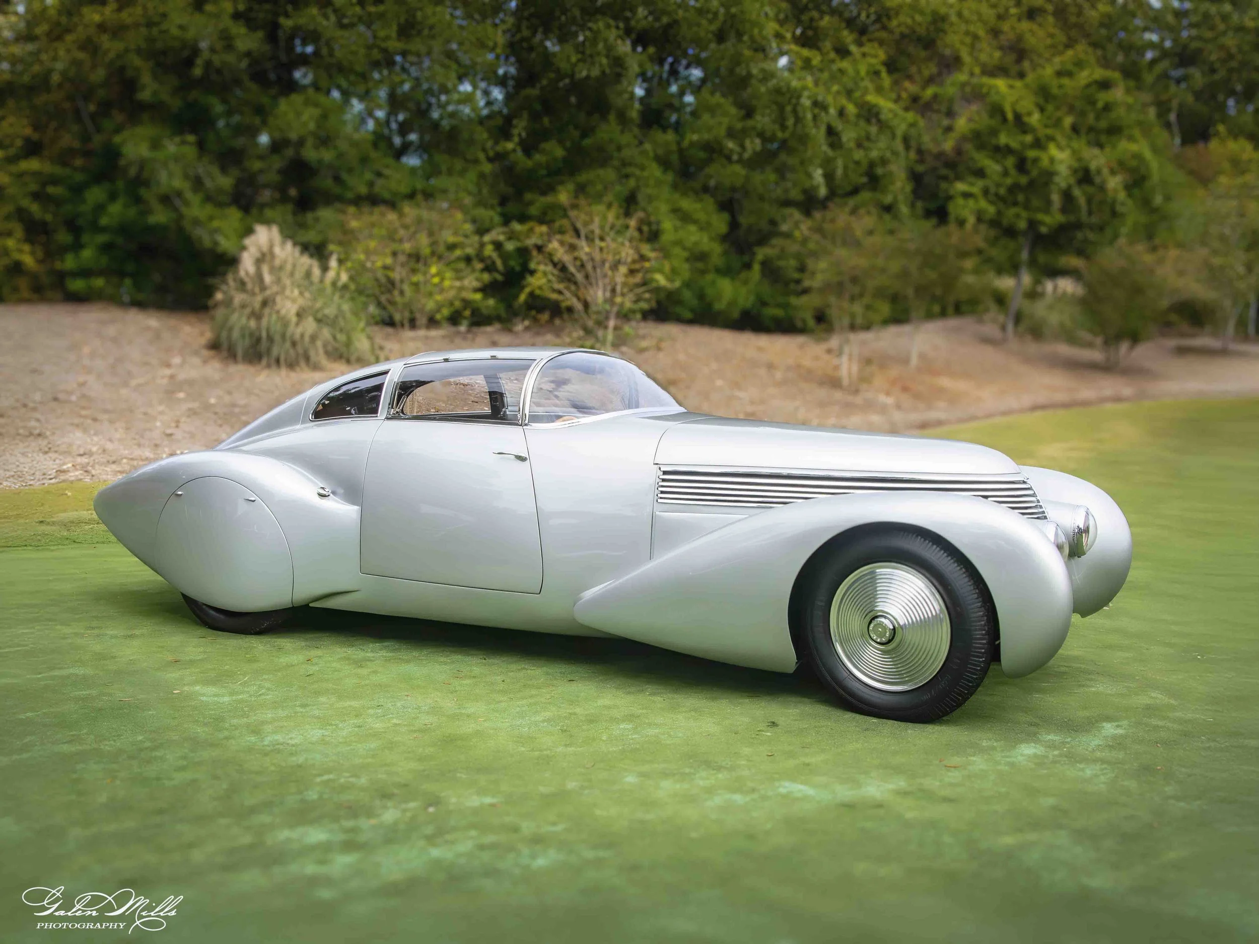 A silver vintage car with a sleek design parked on grass, with trees in the background.