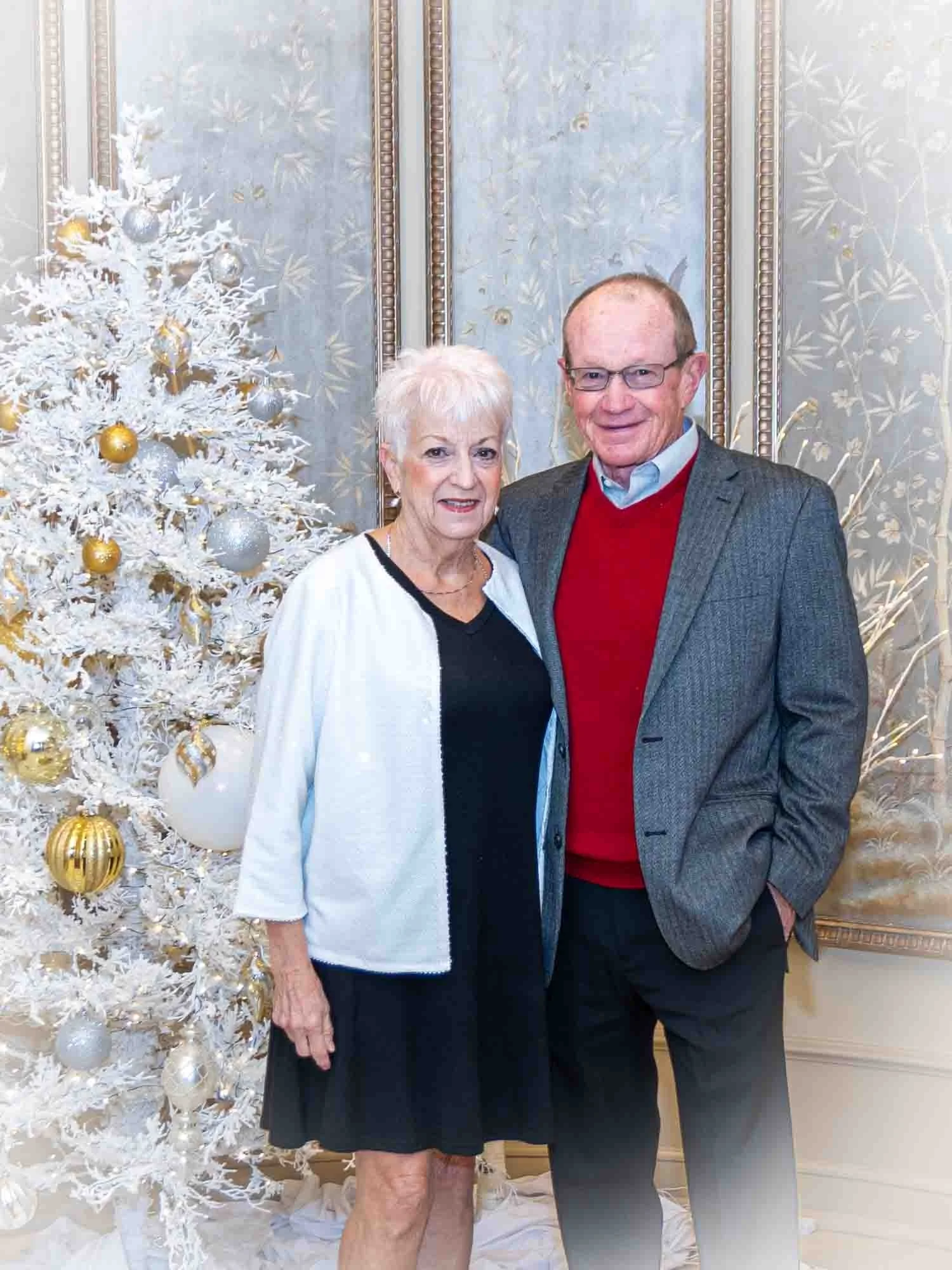 A couple standing next to a white Christmas tree decorated with gold and silver ornaments. The woman is wearing a white cardigan over a black dress, and the man is wearing a gray jacket over a red sweater with a white shirt.
