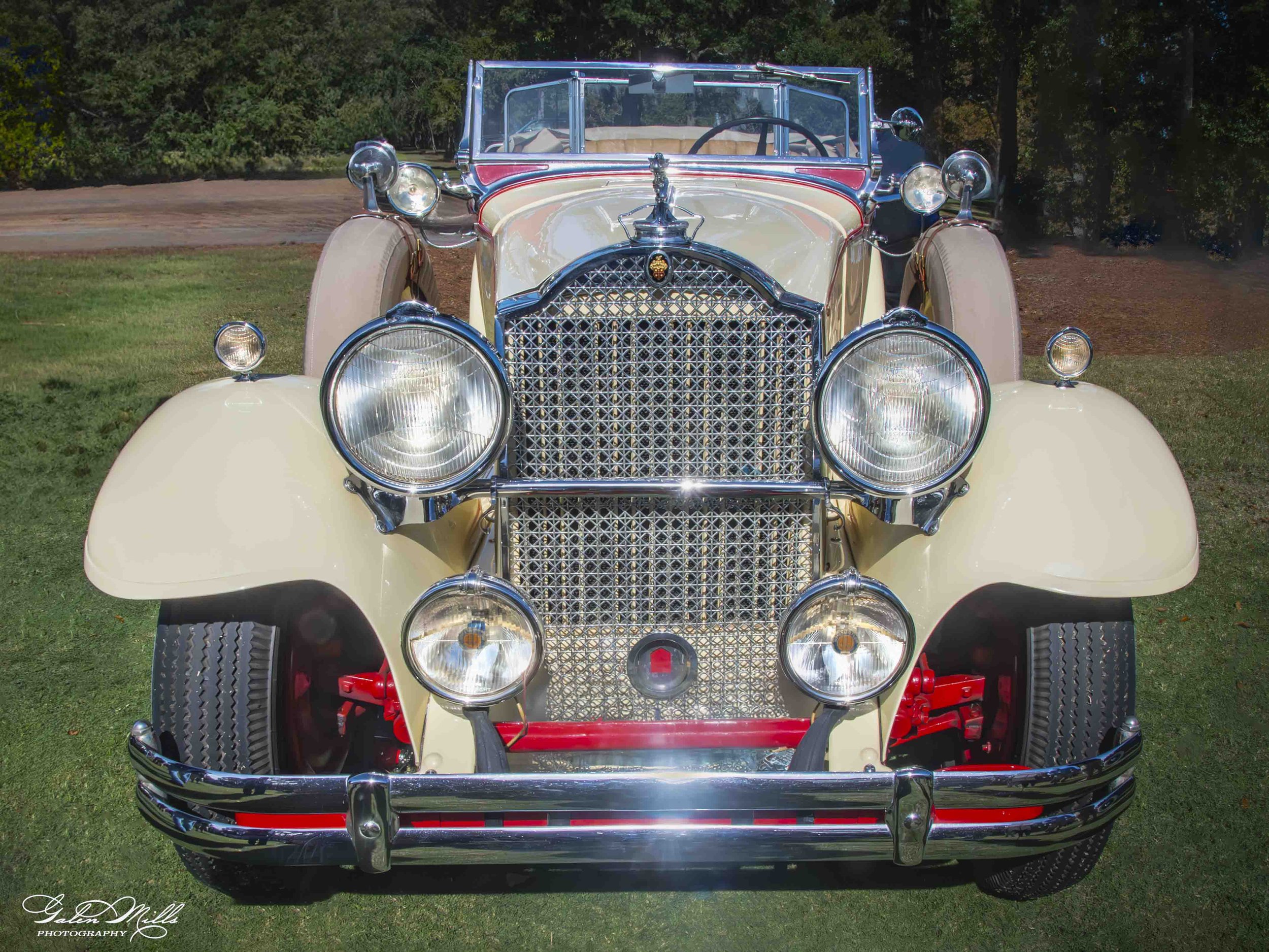 Front view of a vintage cream-colored car with chrome accents, large round headlights, a detailed chrome grille, and a windshield, parked on grass with trees in the background.
