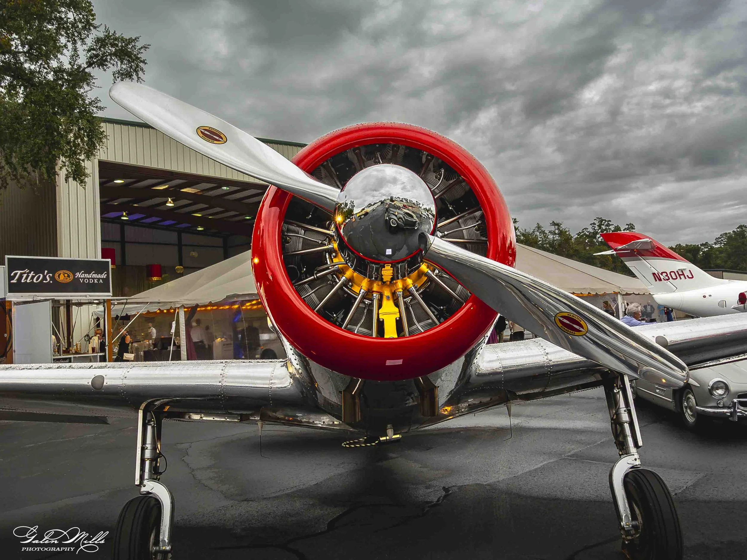 Front view of a vintage propeller airplane parked outdoors during cloudy weather, with hangar and advertising signs in the background.
