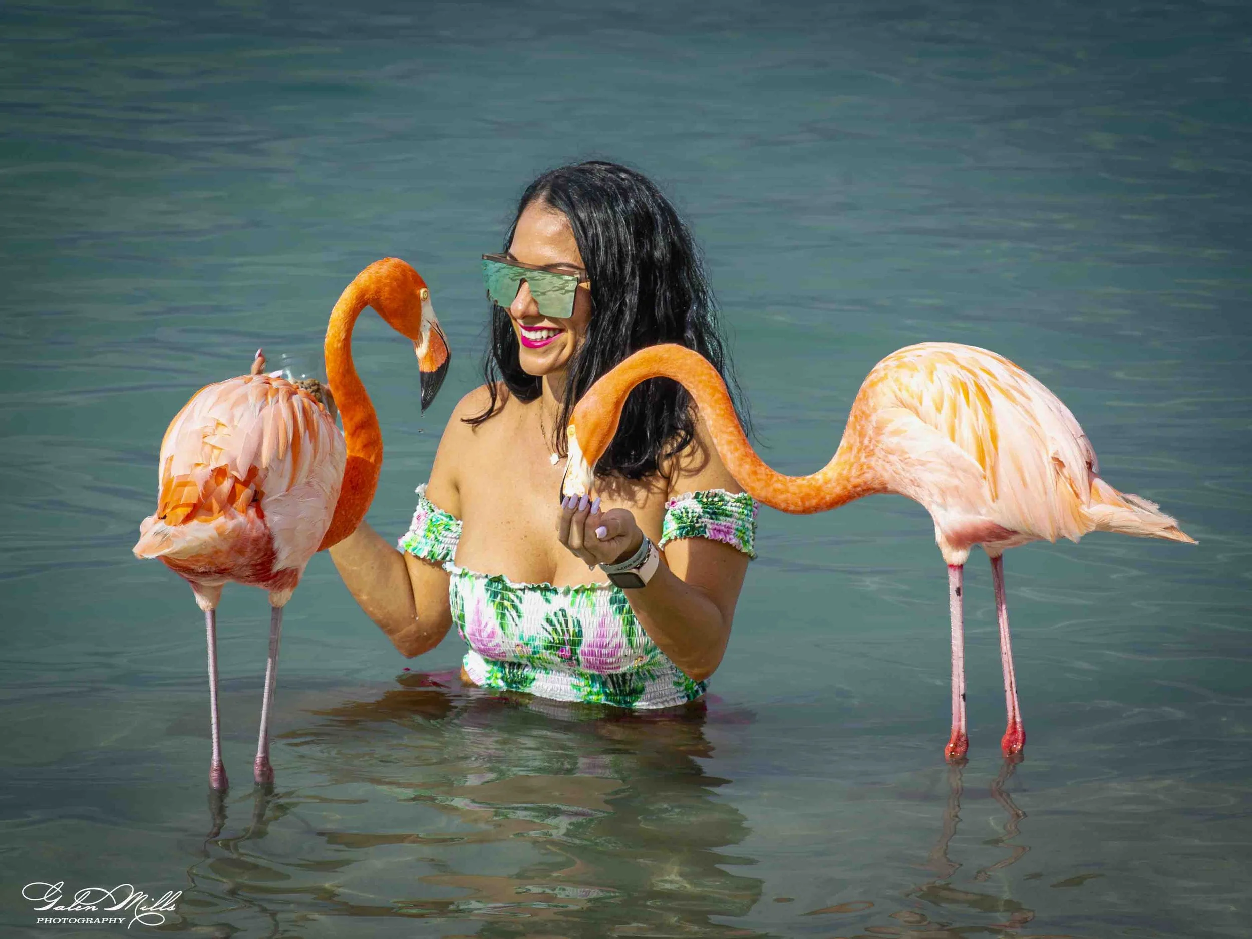 A woman wearing sunglasses and a colorful dress stands in water with three flamingos, smiling and holding a flamingo.