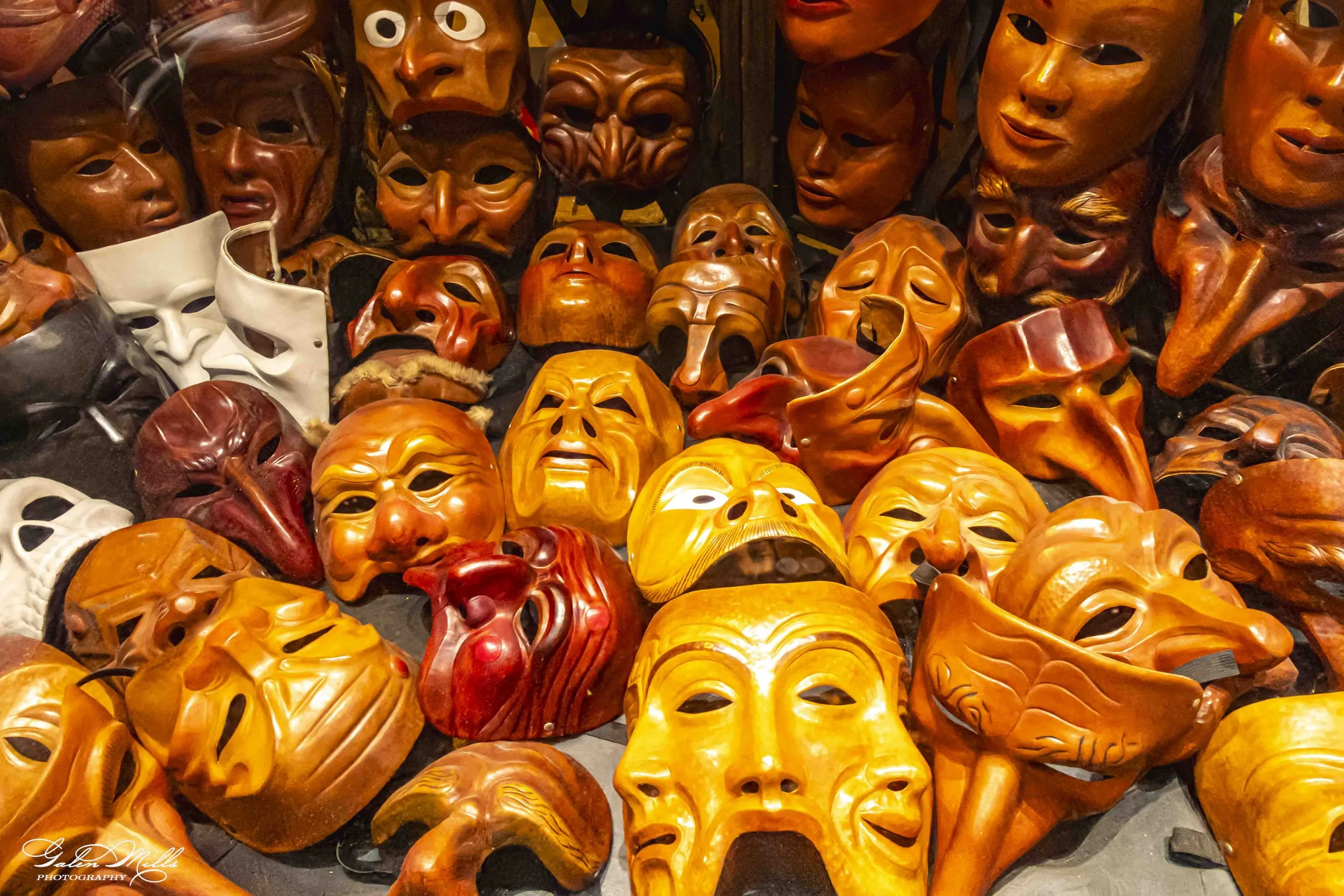 A collection of carved wooden masks in various sizes and expressions, primarily in shades of brown, orange, yellow, and white, displayed in a crowded arrangement.