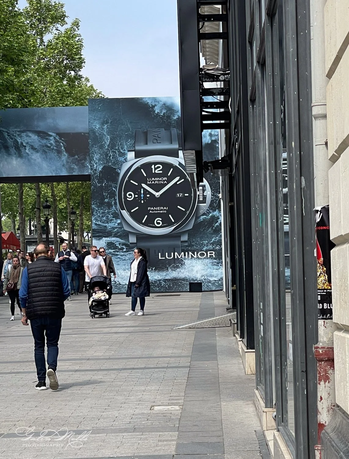 Street scene with large advertisement for Panerai Luminor Marina watch, showing people walking and buildings.
